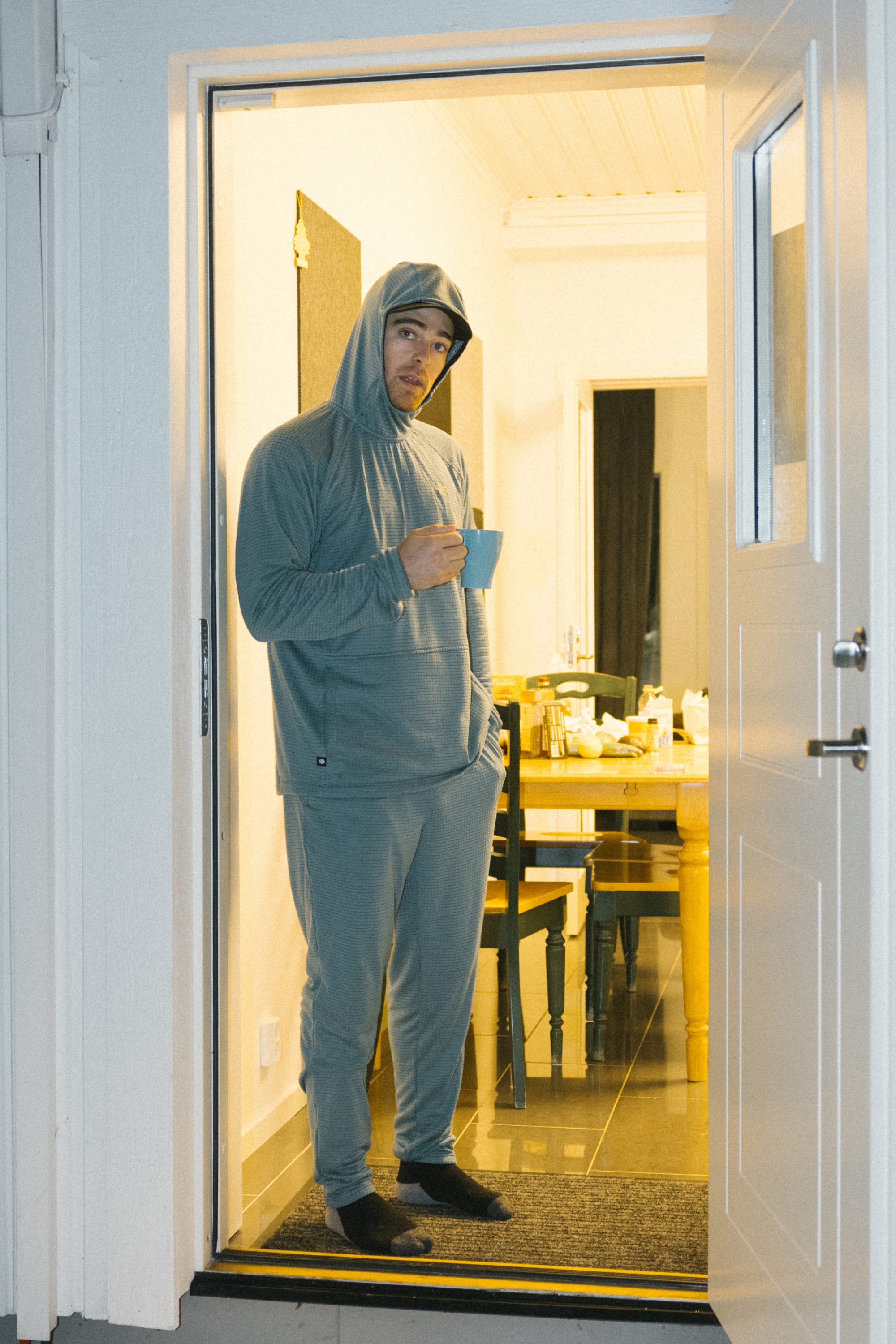 A person wearing the 686 Men’s Mission Grid™ Raglan Hoody in gray stands in a doorway, one hand in their pocket and holding a blue mug, gazing at the camera. The warmly lit indoor room can be seen behind them.