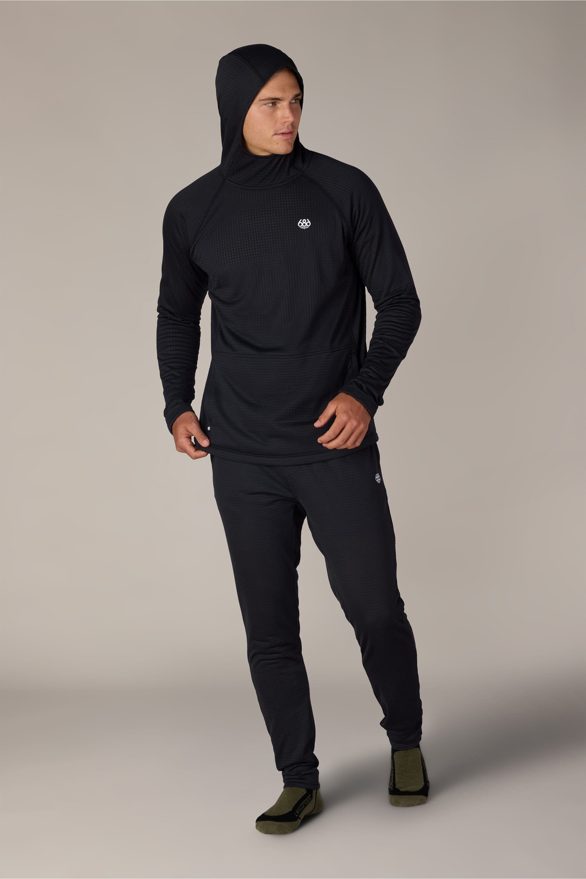 A man wearing a black hooded long-sleeve shirt and the 686 Men’s Mission Grid™ Pant from 686 stands against a plain background, looking to the side with one hand near his hip and the other relaxed by his side. He wears olive slippers.