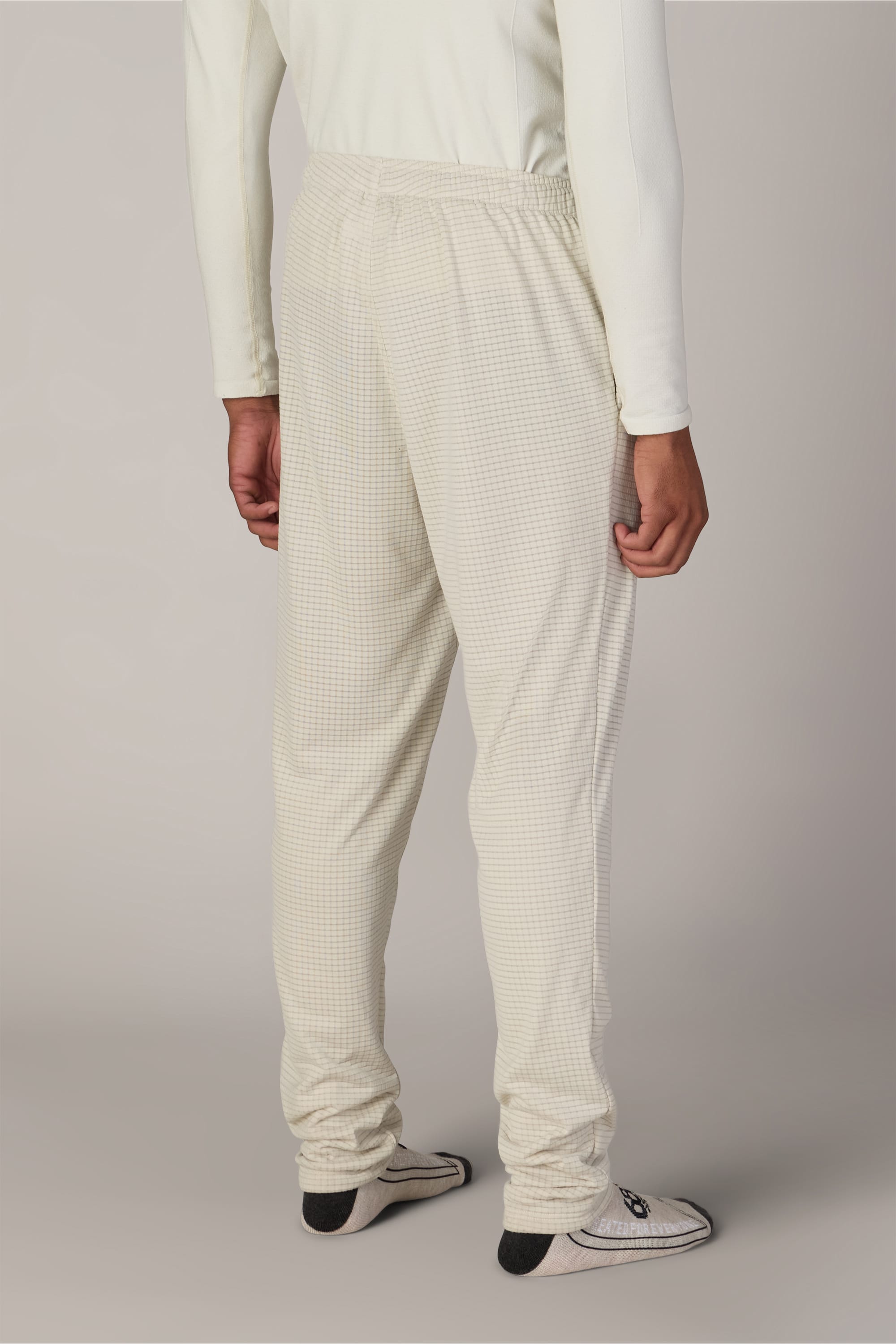 A person stands facing away, wearing a long-sleeve light shirt, the 686 Men’s Mission Grid™ Pant by 686 in a light-colored checkered pattern, and white socks with black non-slip grips. The background is plain and neutral.