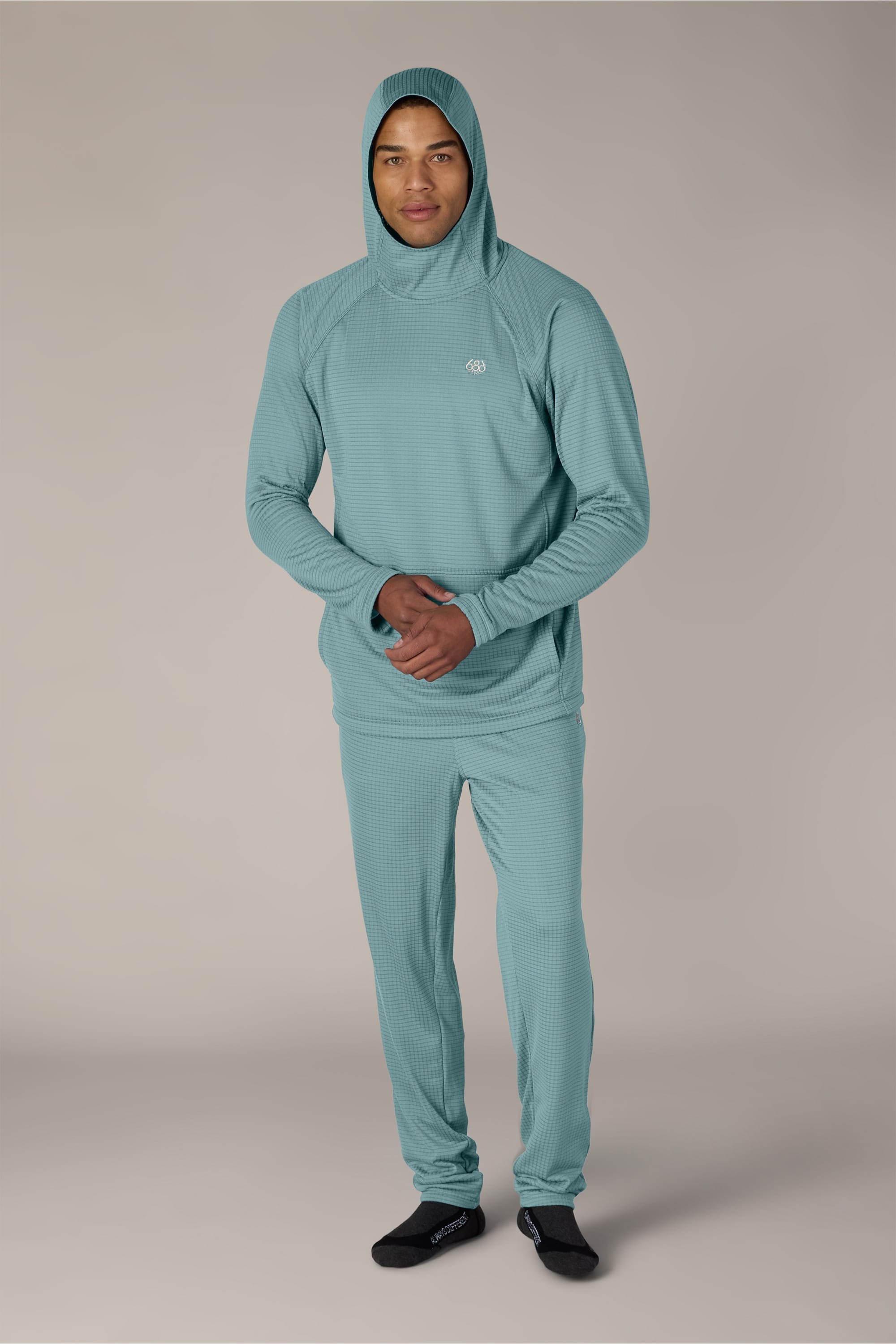 A person stands facing forward, wearing a light teal hooded sweatshirt and 686 Men’s Mission Grid™ Pant by 686, paired with black socks, against a plain light background.