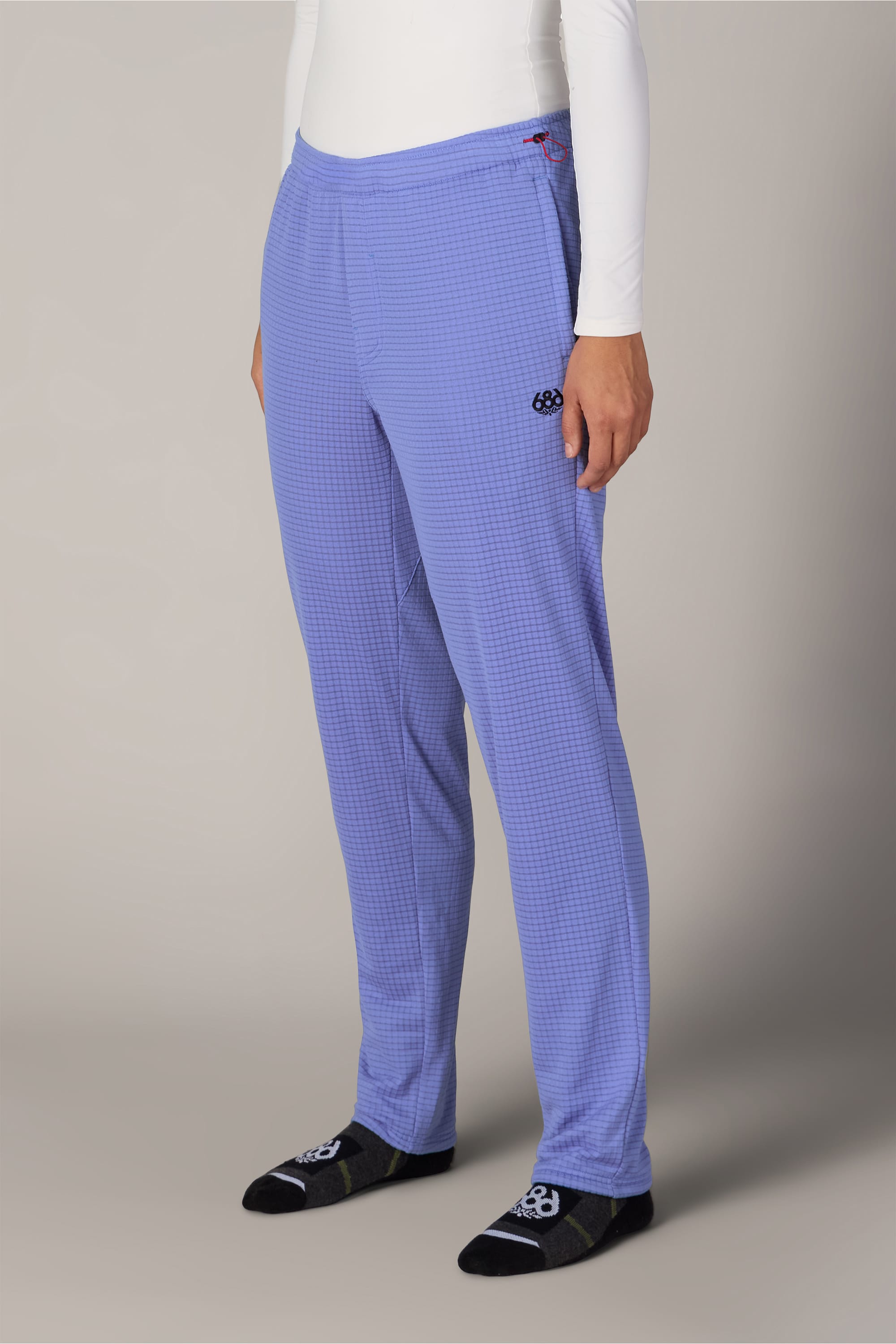 A person is wearing a white long-sleeve shirt, 686 Women’s Mission Grid™ Pant in blue checkered knit with a small black 686 logo, and black socks with light gray patterns, standing against a plain light background.