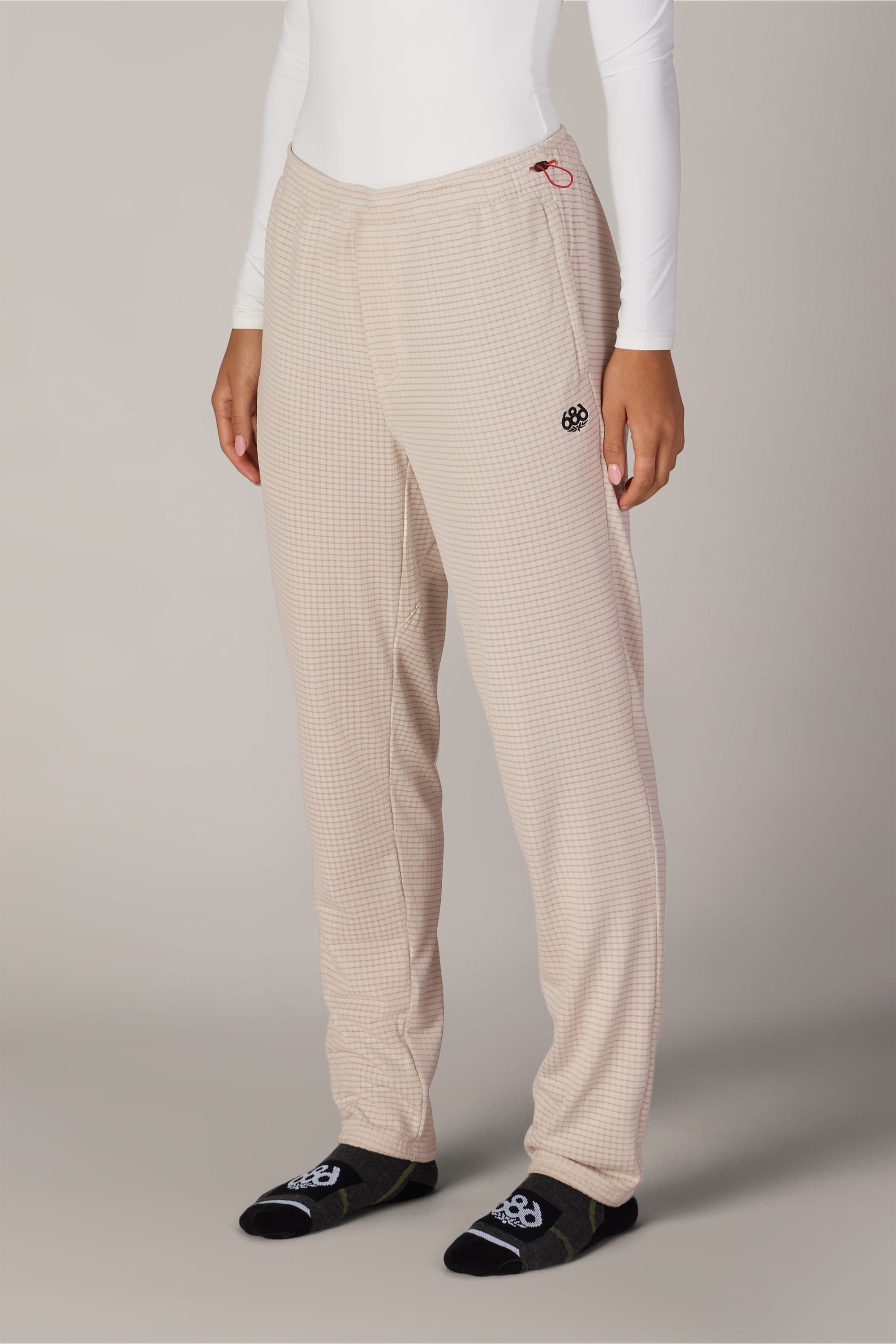 A person is shown from the waist down, wearing the 686 Women’s Mission Grid™ Pant by 686 in a beige checkered pattern, paired with a white long-sleeve top and black socks featuring white designs, standing against a plain background.