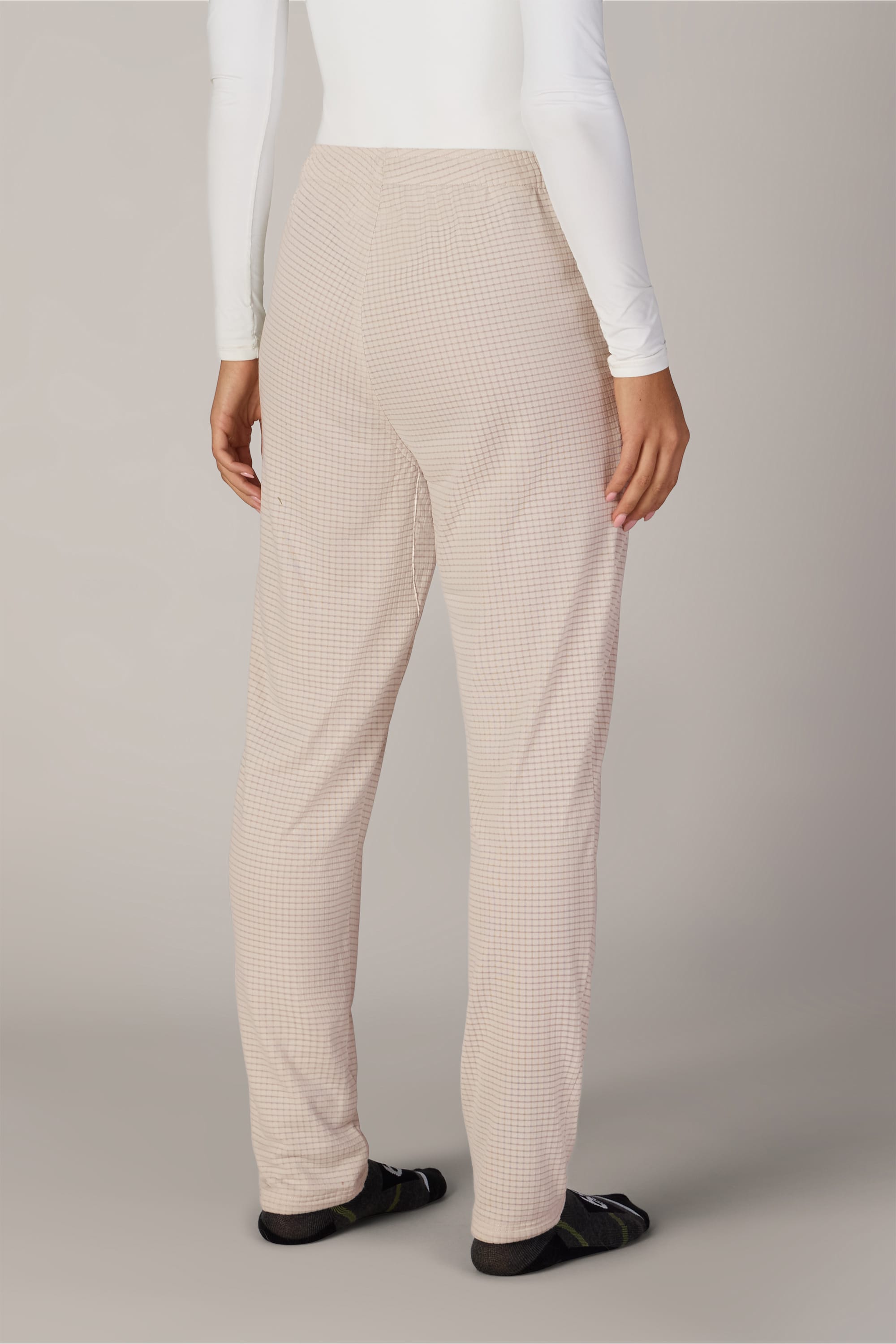 A person stands facing away, wearing 686 Women’s Mission Grid™ Pants by 686 in a beige checkered pattern, paired with a white long-sleeve shirt and black socks. The background is plain and light-colored.