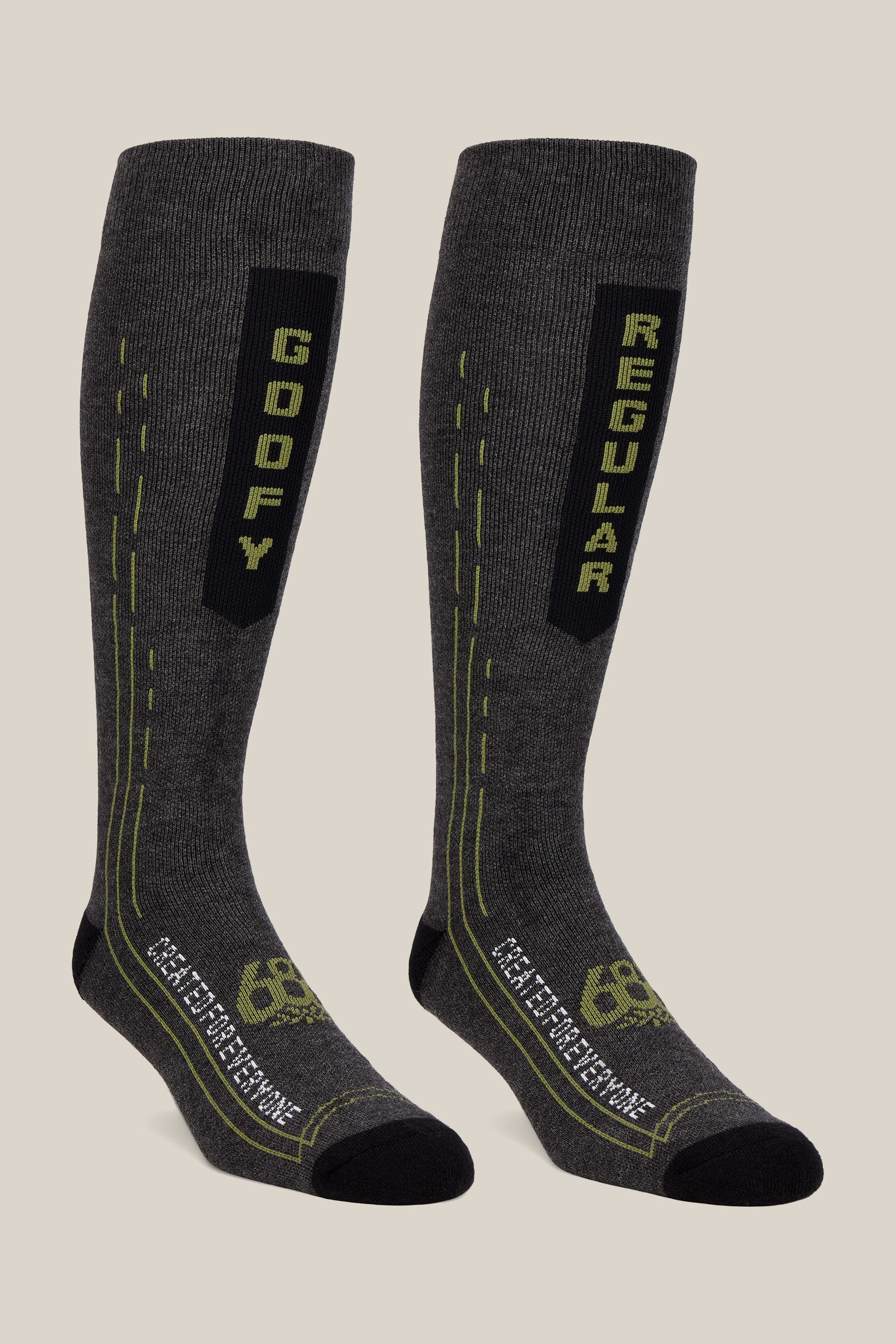 A pair from the 686 Men’s Merino Balance Sock 2-Pack features tall, dark gray performance socks with black toes and heels; one displays “GOOFY” in green text while the other reads “REGULAR.” Both socks include green lines and patterns, subtle 686 branding near the toes, and are designed for optimal comfort and style.