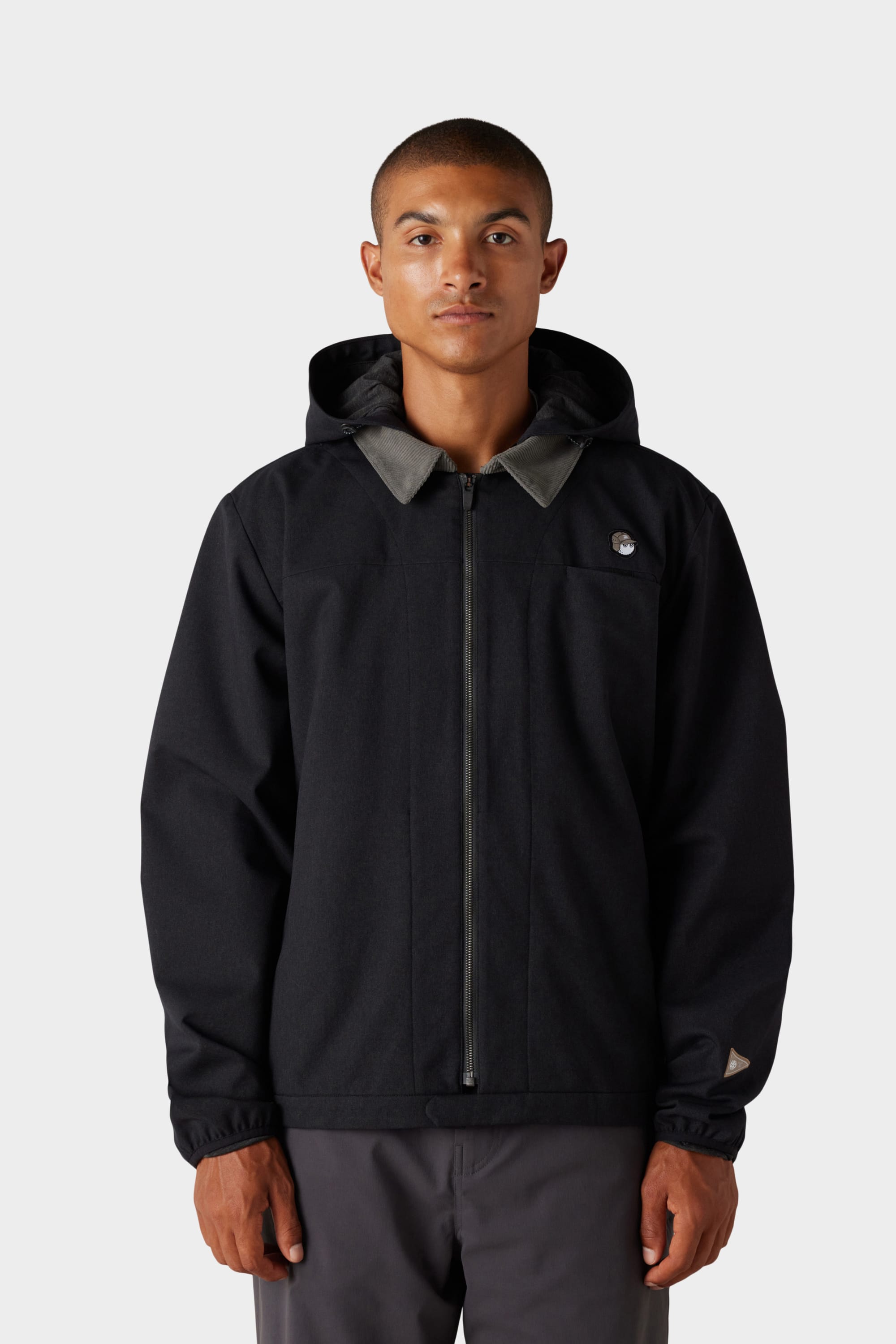 A person faces forward against a light grey background, wearing the 686 x Malbon Mens Caddie Performance Jacket by 686—a black hooded jacket with a front zipper and a small circular logo on the left chest.