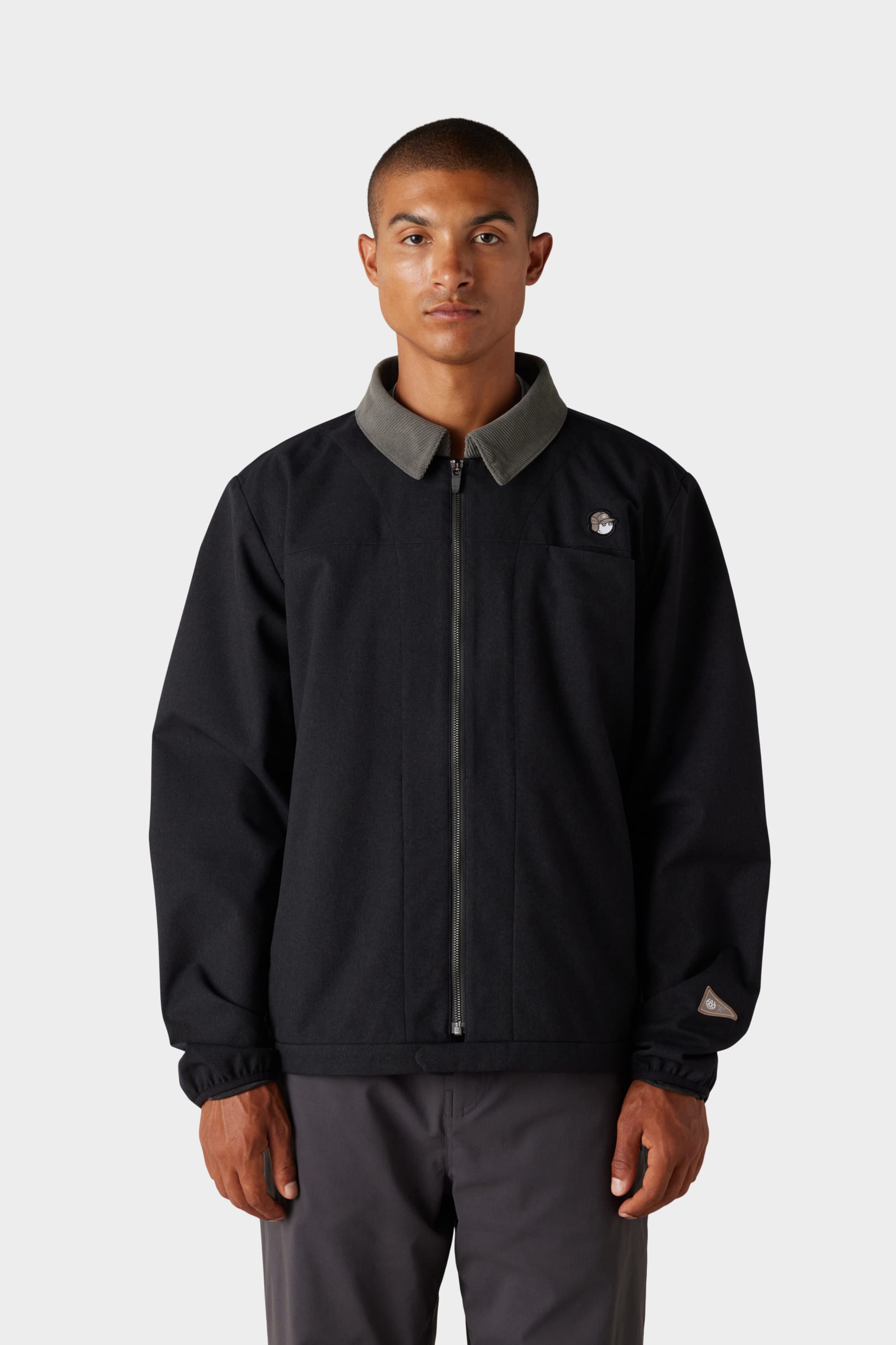 A person stands facing forward, wearing the 686 x Malbon Mens Caddie Performance Jacket by 686, which is black with a grey collar, paired with dark pants against a plain light grey background.