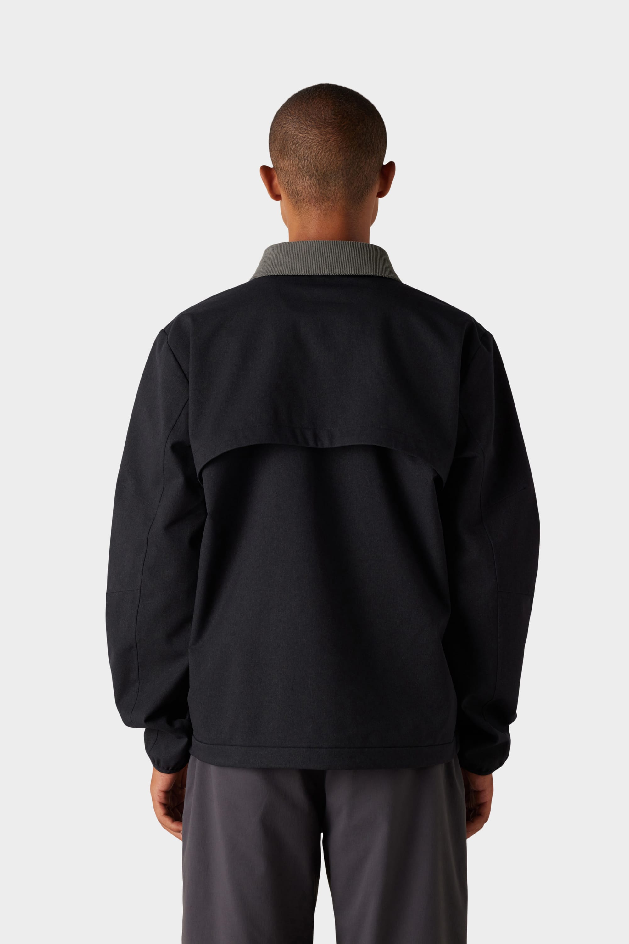 A person with short hair faces away, wearing the 686 x Malbon Mens Caddie Performance Jacket by 686—black with a grey collar—and dark pants, set against a plain light background.