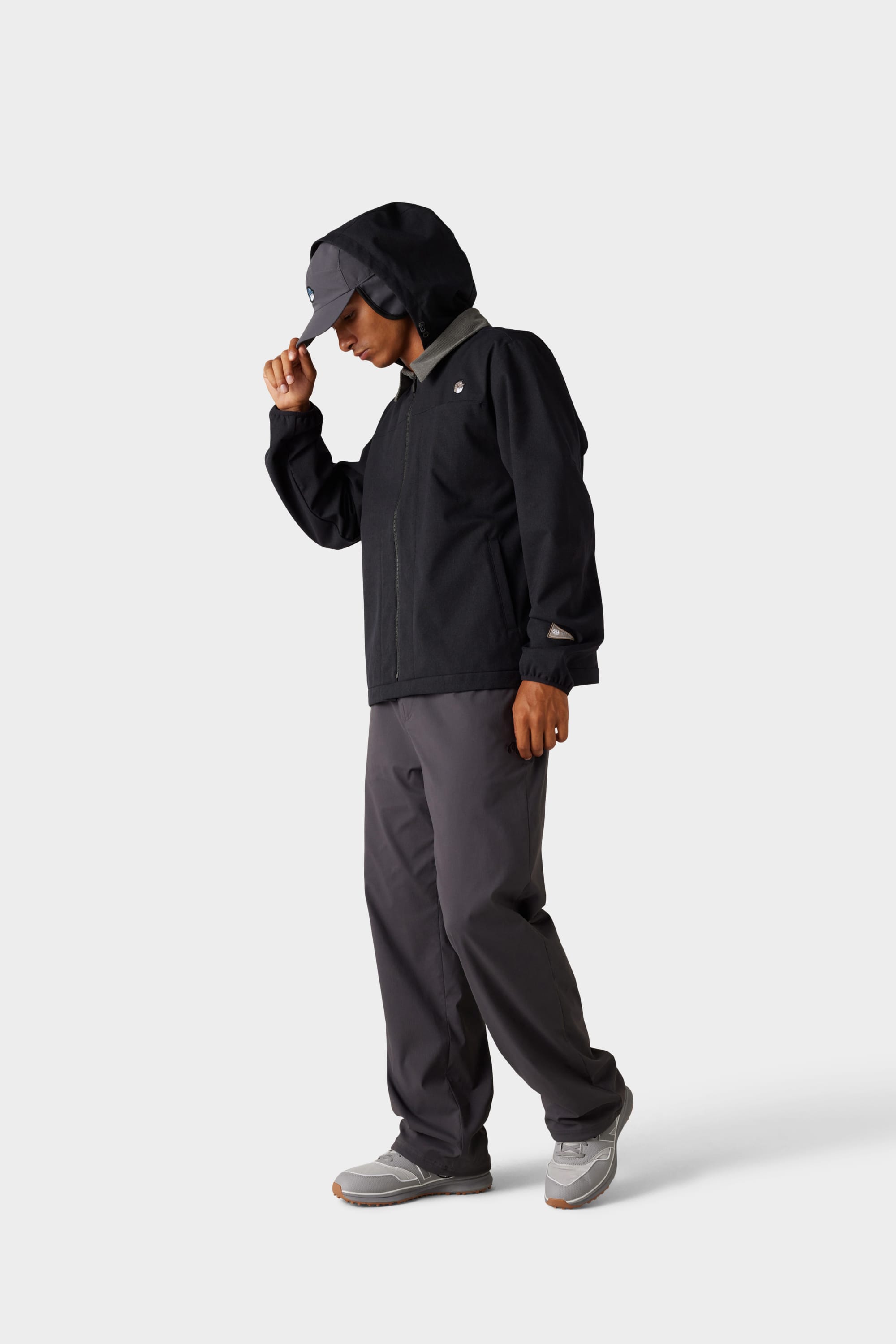 A person wearing the 686 x Malbon Mens Caddie Performance Jacket by 686, along with grey pants, grey sneakers, and a grey cap stands against a plain white background, looking down and adjusting their cap.