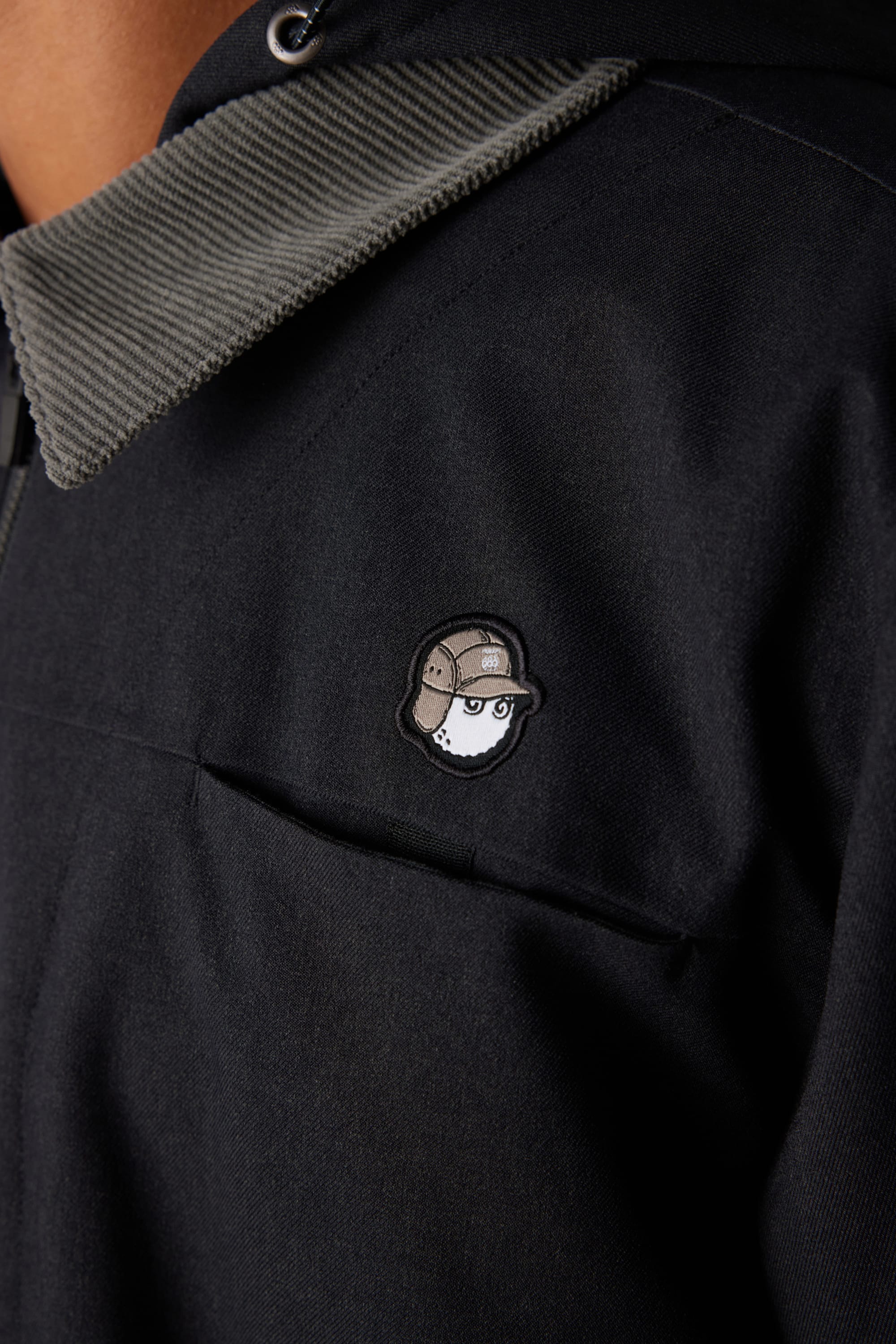 Close-up of a person wearing the 686 x Malbon Mens Caddie Performance Jacket by 686, featuring a dark design, ribbed grey collar, and an embroidered cartoon face with a detective hat on the chest.