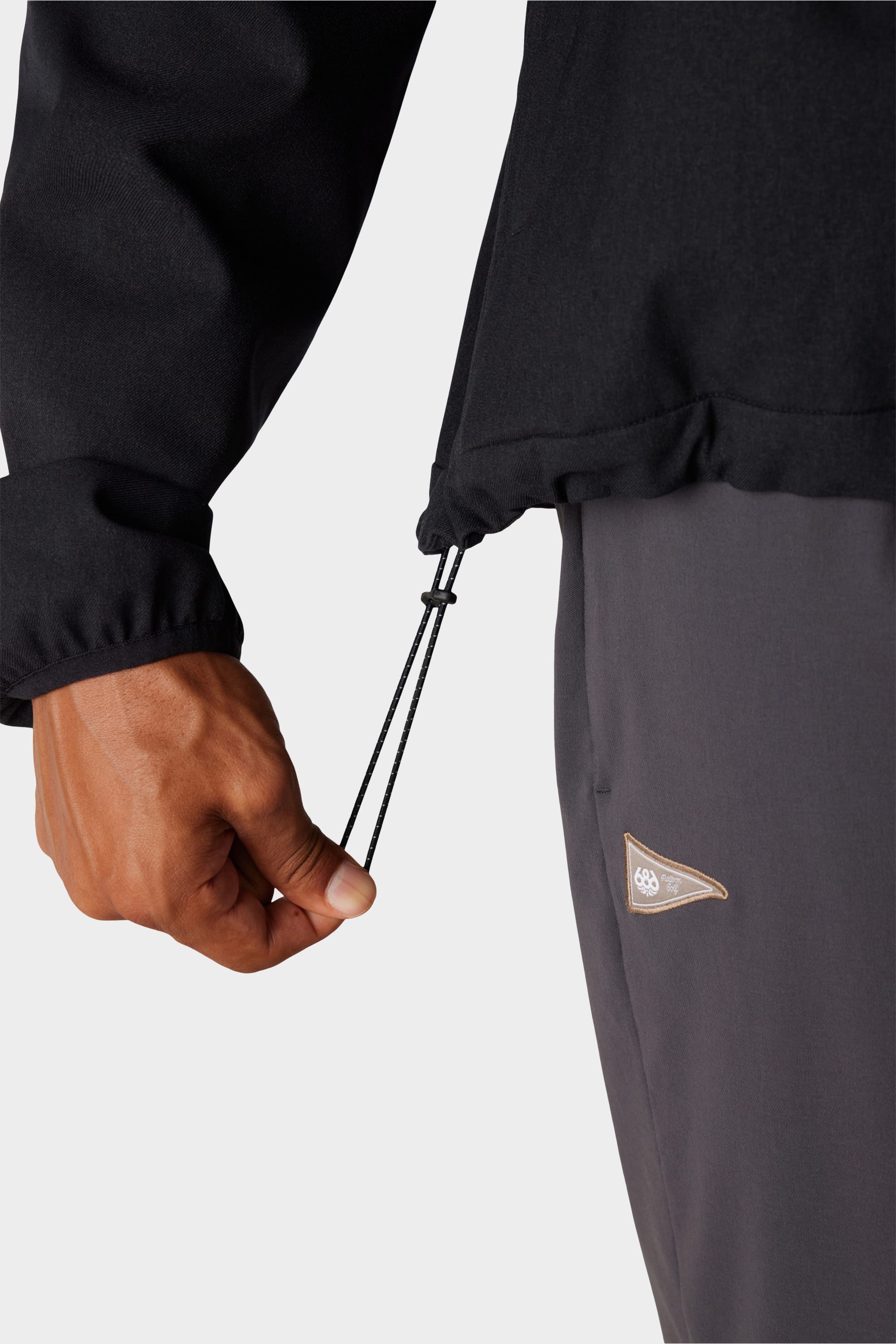 A person adjusts the drawstring hem of a 686 x Malbon Mens Caddie Performance Jacket in black, paired with dark grey pants featuring a small triangular logo, set against a plain light background.