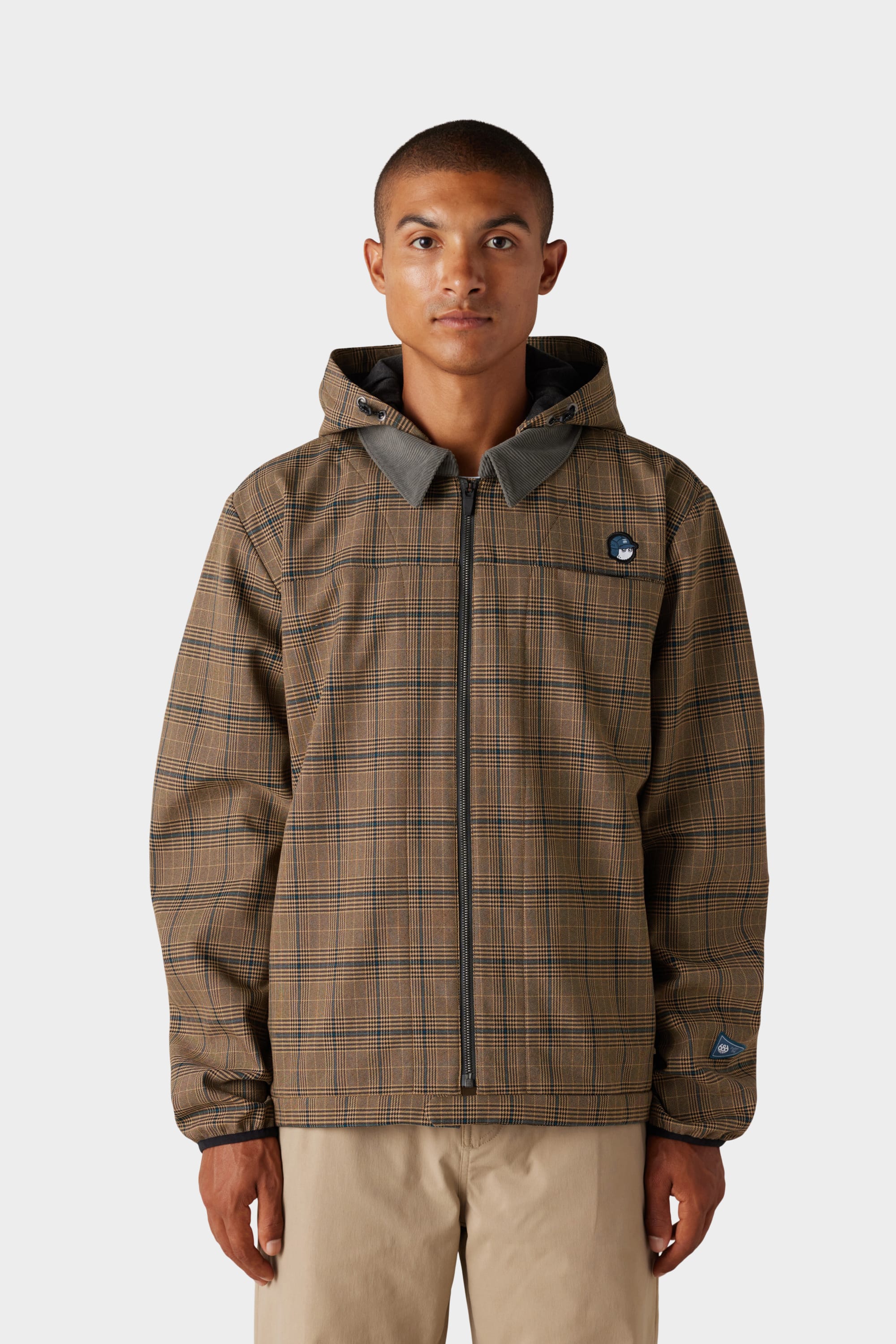 A person faces forward against a plain light background wearing the 686 x Malbon Mens Caddie Performance Jacket by 686, featuring brown plaid, a hood, and front zipper, paired with khaki pants.