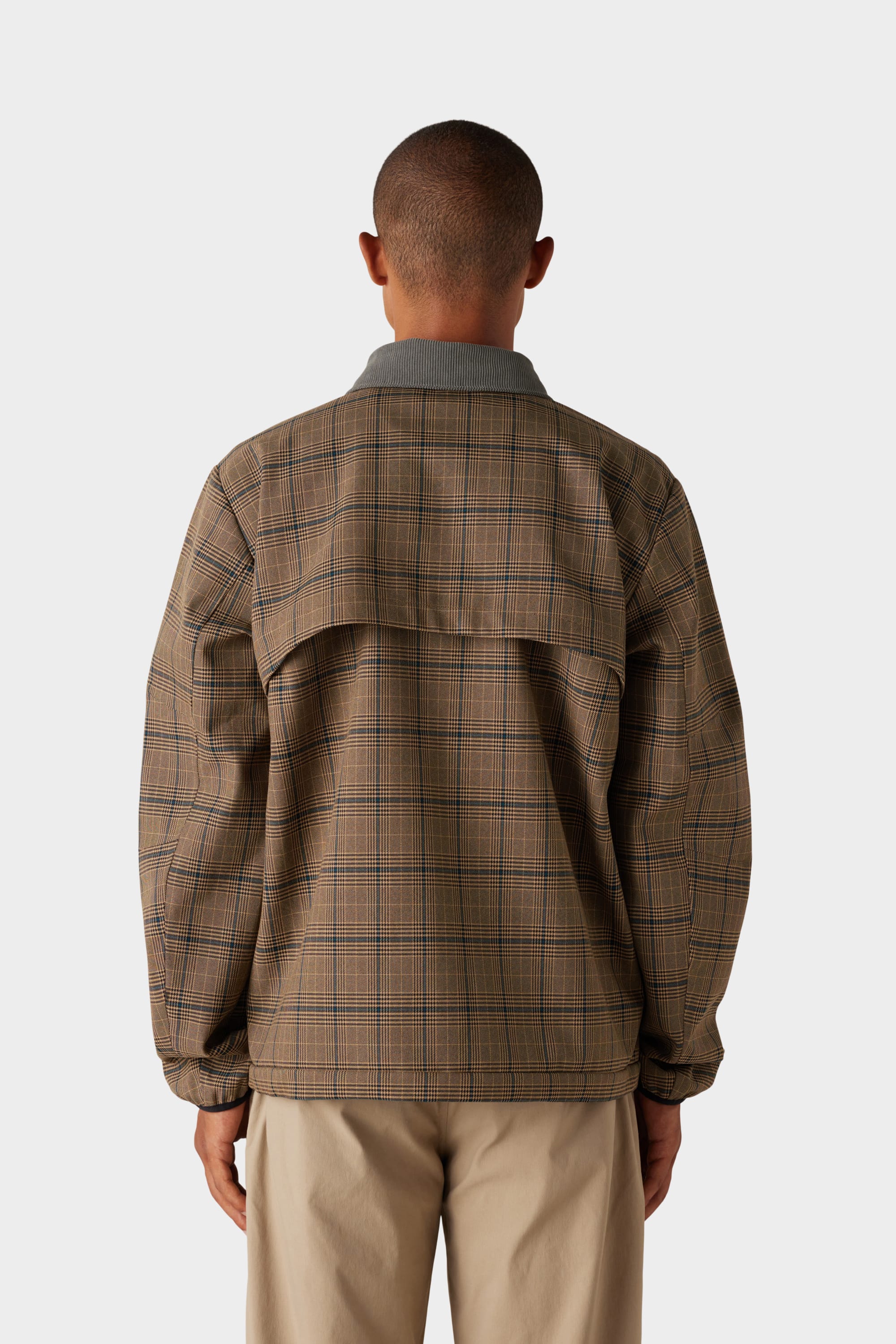 A person stands facing away, wearing the 686 x Malbon Mens Caddie Performance Jacket by 686, featuring a brown plaid design with a back yoke detail, paired with light-colored pants. The background is plain and light grey.