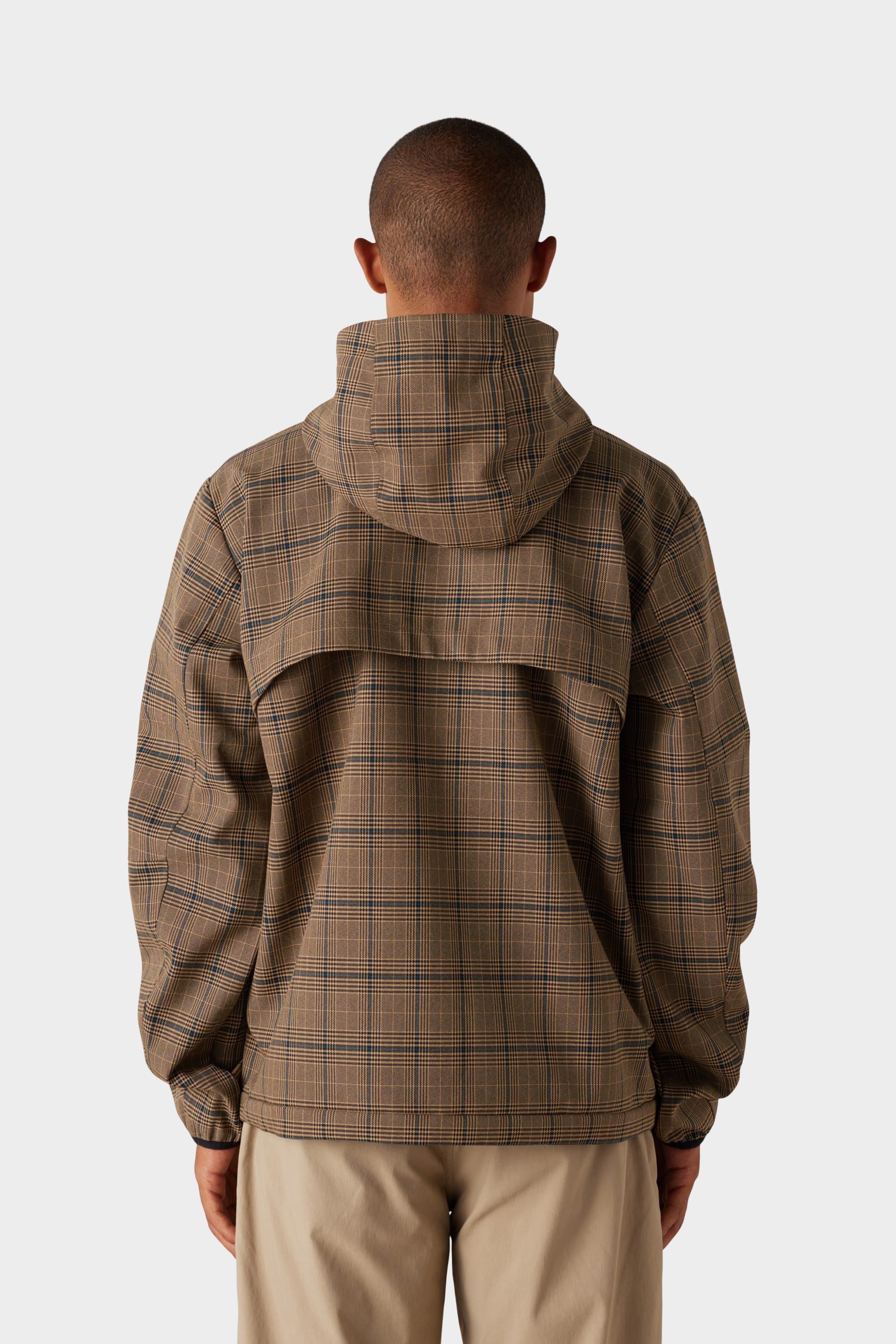 A person faces away, wearing the 686 x Malbon Mens Caddie Performance Jacket by 686 in brown plaid with a vented back, paired with beige pants. The background is plain white.
