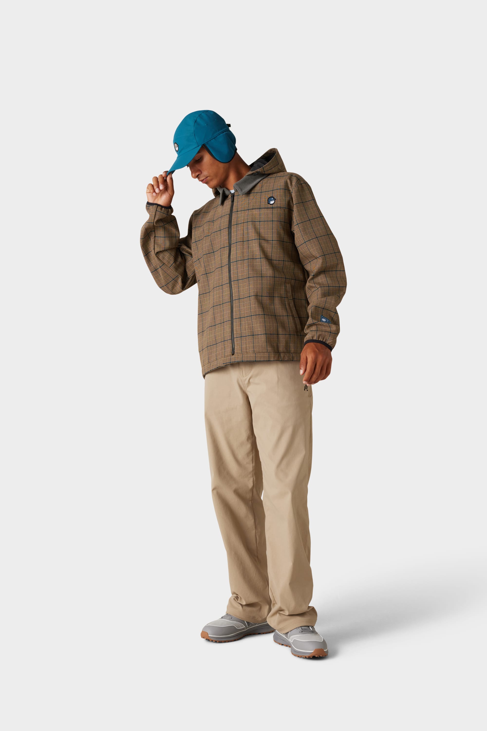 A person wearing a blue cap, the 686 x Malbon Mens Caddie Performance Jacket by 686, beige pants, and grey sneakers stands against a plain white background, looking downward and touching the brim of the cap.