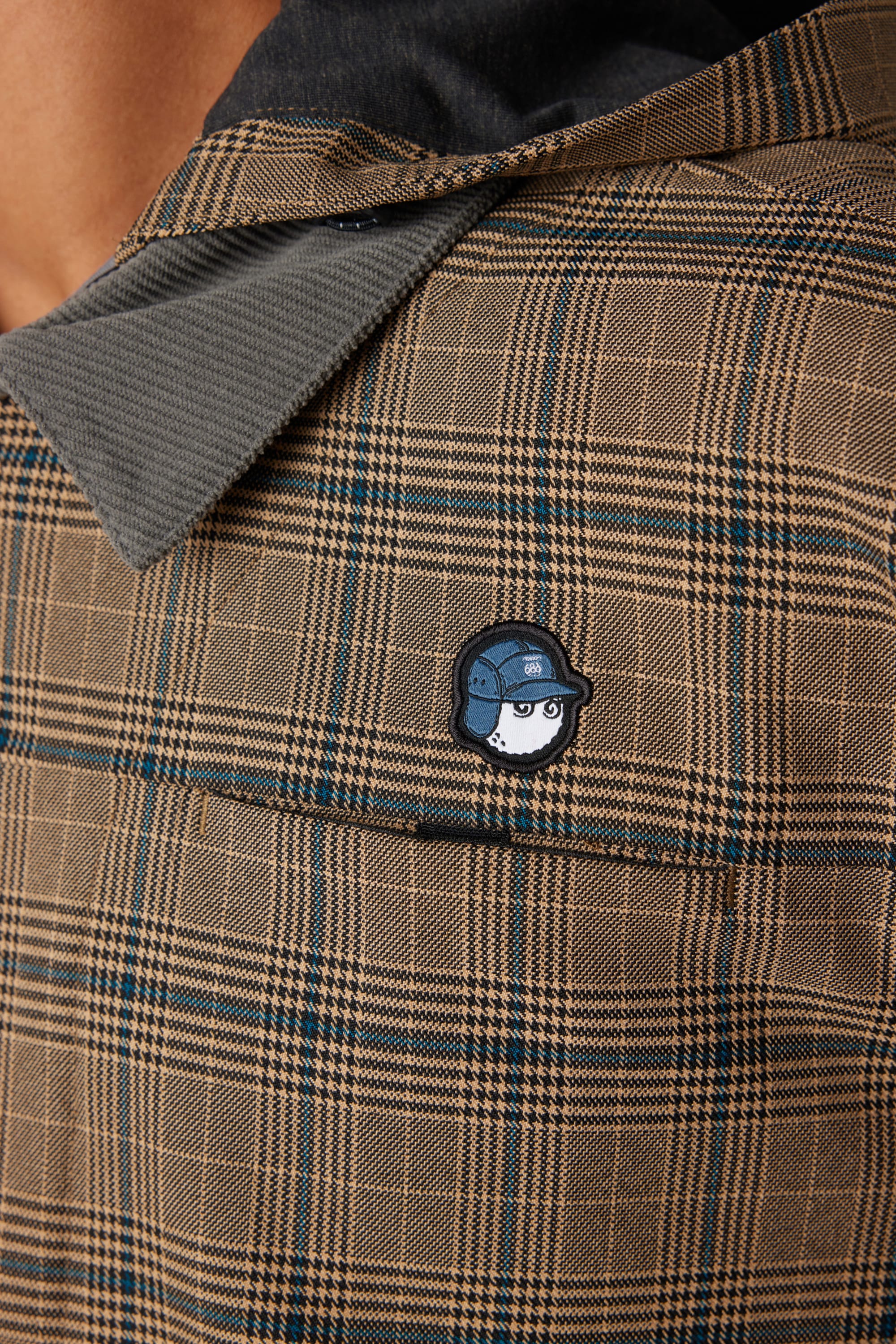A close-up of the 686 x Malbon Mens Caddie Performance Jacket features a chest patch with a cartoon face wearing glasses and a helmet, along with the jacket collar and part of the wearer’s neck.