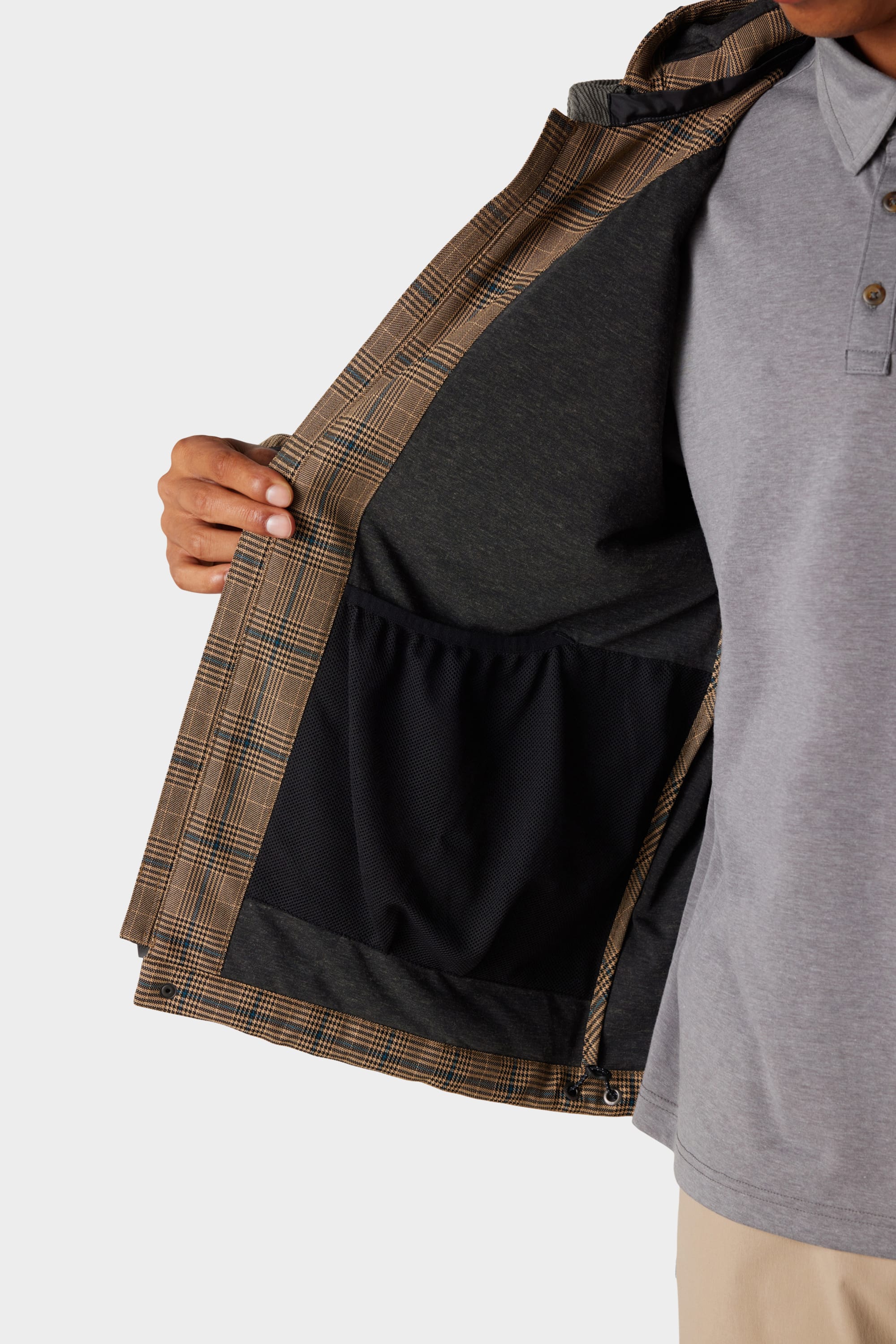A person holds open the 686 x Malbon Mens Caddie Performance Jacket by 686, revealing its black inner lining and interior pocket. The jacket features a brown plaid exterior; the wearer’s face is not visible.
