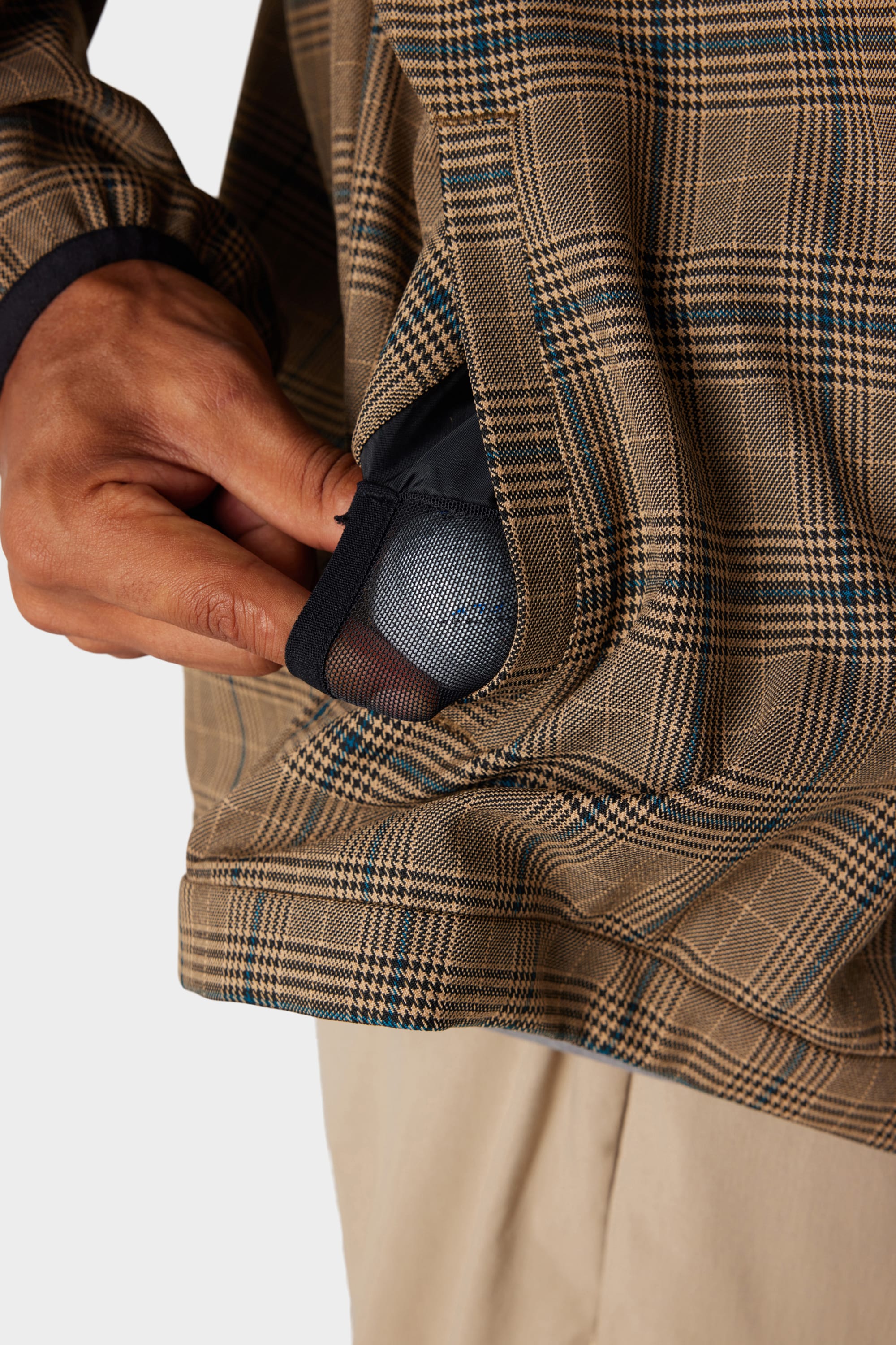 A person’s hand slips a small, round mesh drawstring pouch into the side pocket of the 686 x Malbon Mens Caddie Performance Jacket, which features a brown and blue plaid design. The individual is dressed in light khaki pants.