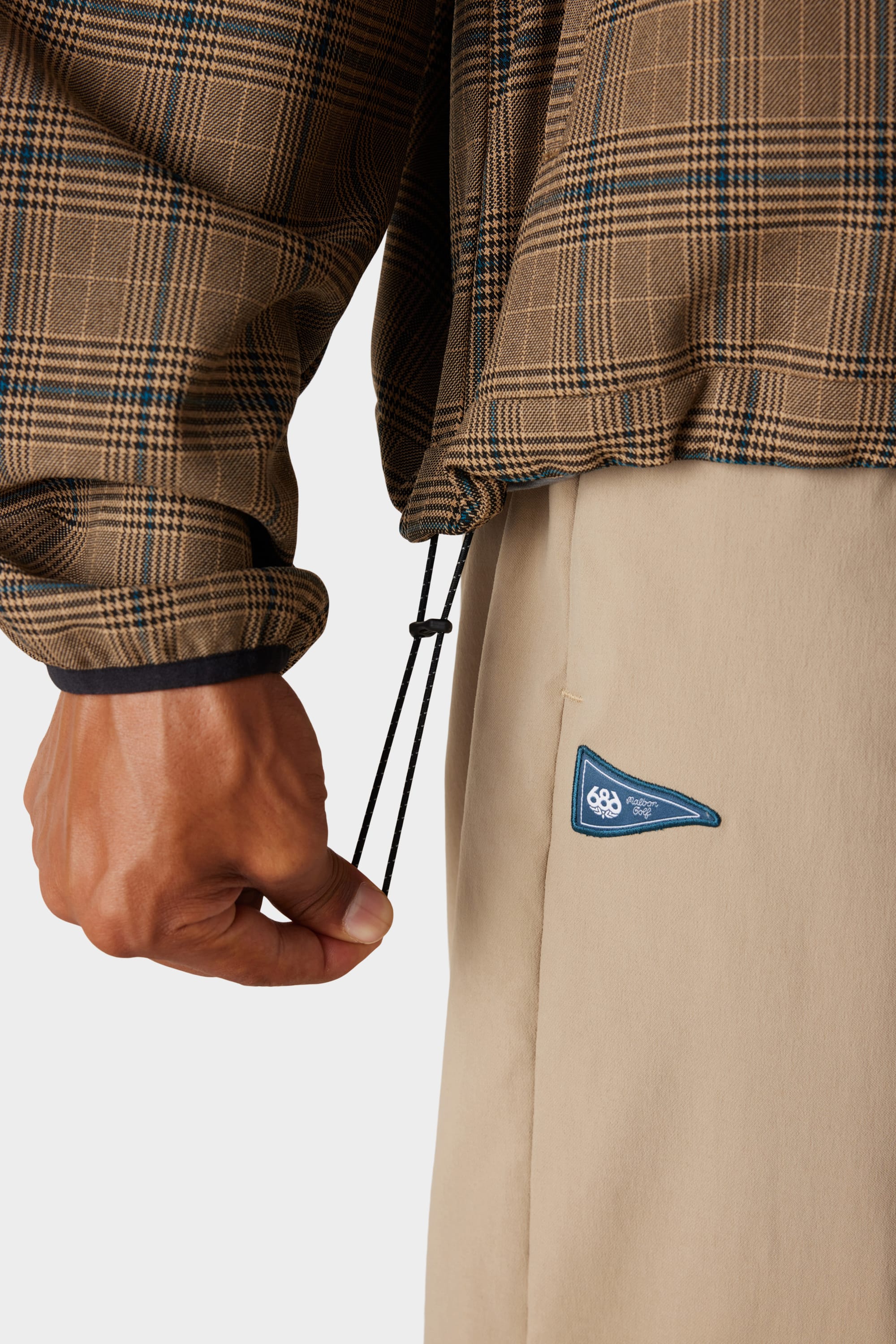 A person’s left hand pulls a black drawstring on the 686 x Malbon Mens Caddie Performance Jacket, a brown plaid piece by 686, styled with beige pants featuring a small blue and white patch on the pocket.