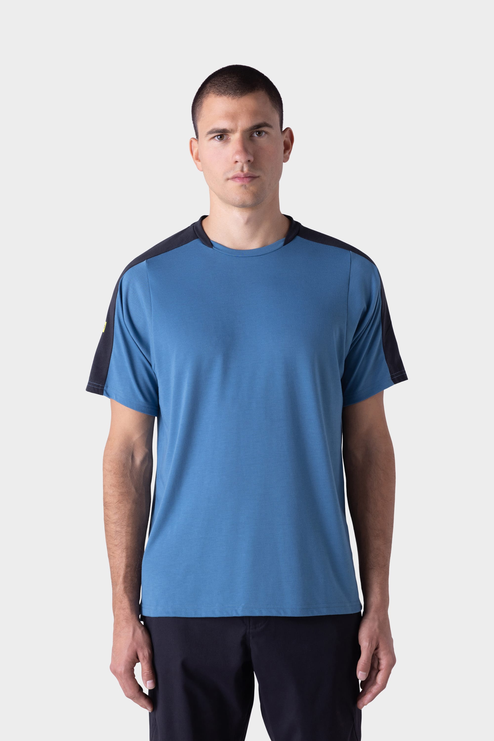 A man with short hair wears the 686 Mens Rival Short Sleeve Bike Jersey by 686, featuring blue with black sleeves, and dark pants, as he stands facing forward against a plain light grey background.