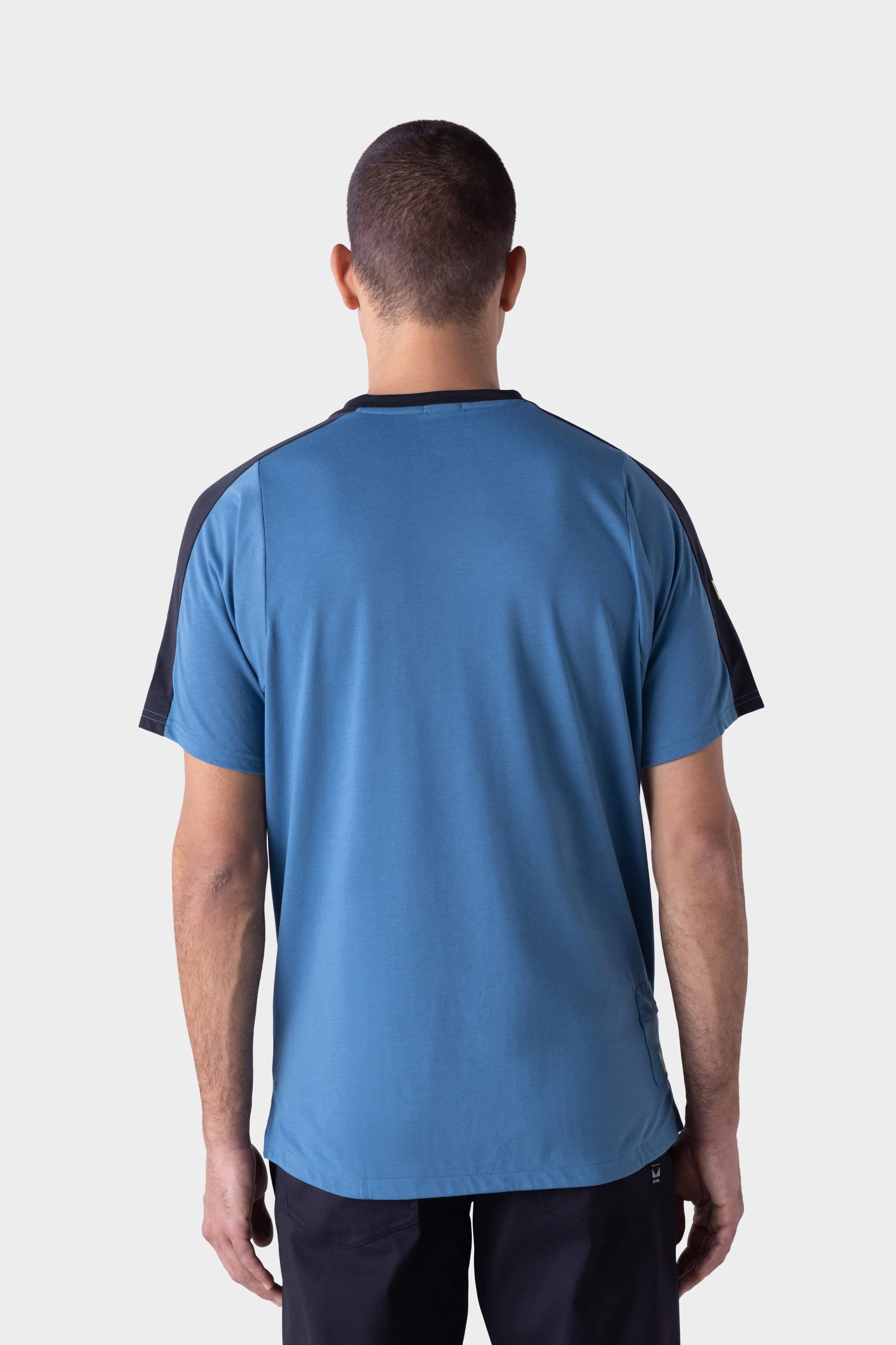 A man with short hair stands facing away, wearing the 686 Mens Rival Short Sleeve Bike Jersey in blue with black shoulder panels and black pants against a plain light background.