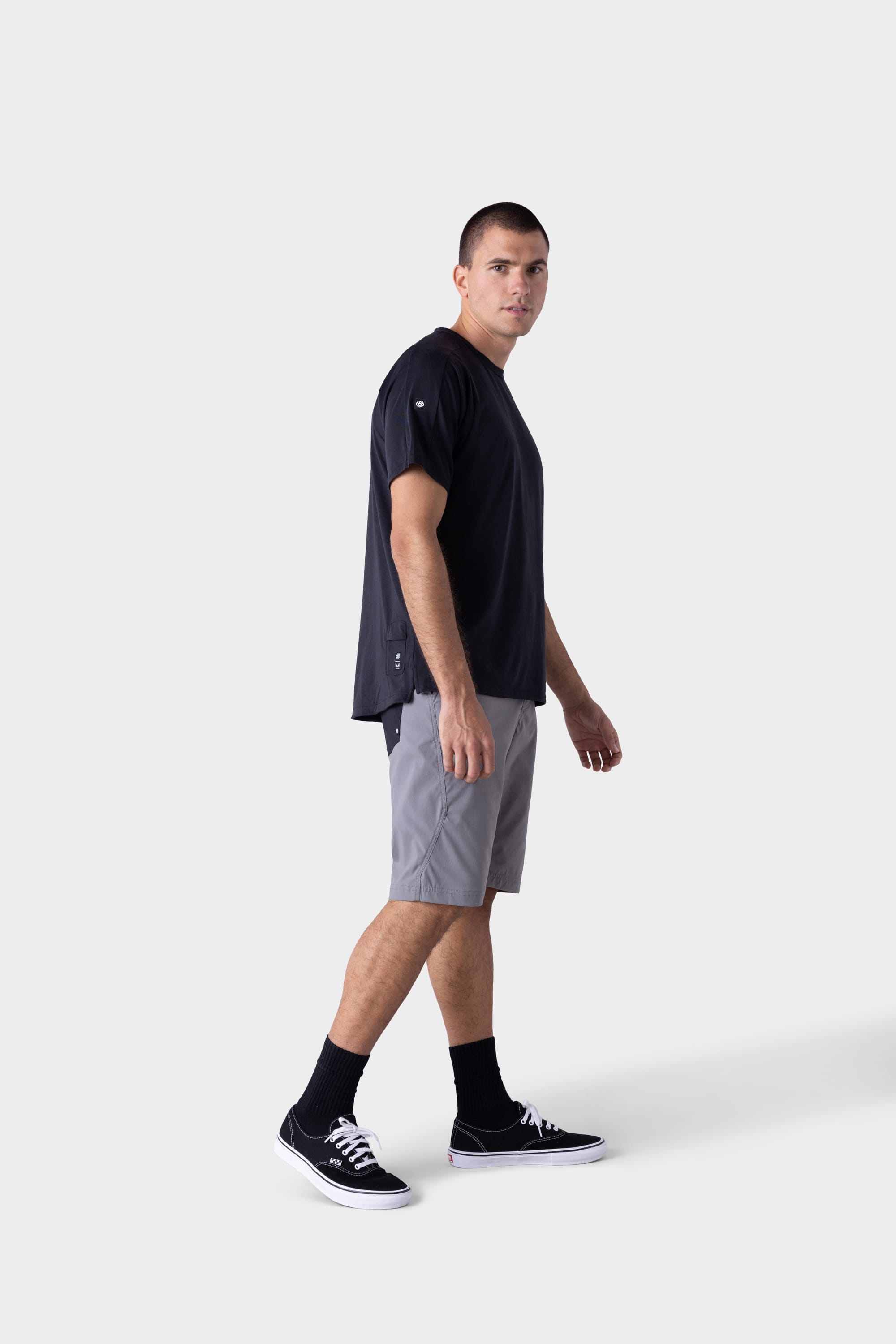 A man wearing the 686 Mens Rival Short Sleeve Bike Jersey stands against a plain light background, paired with light grey shorts, black socks, and black sneakers, looking slightly to the side.