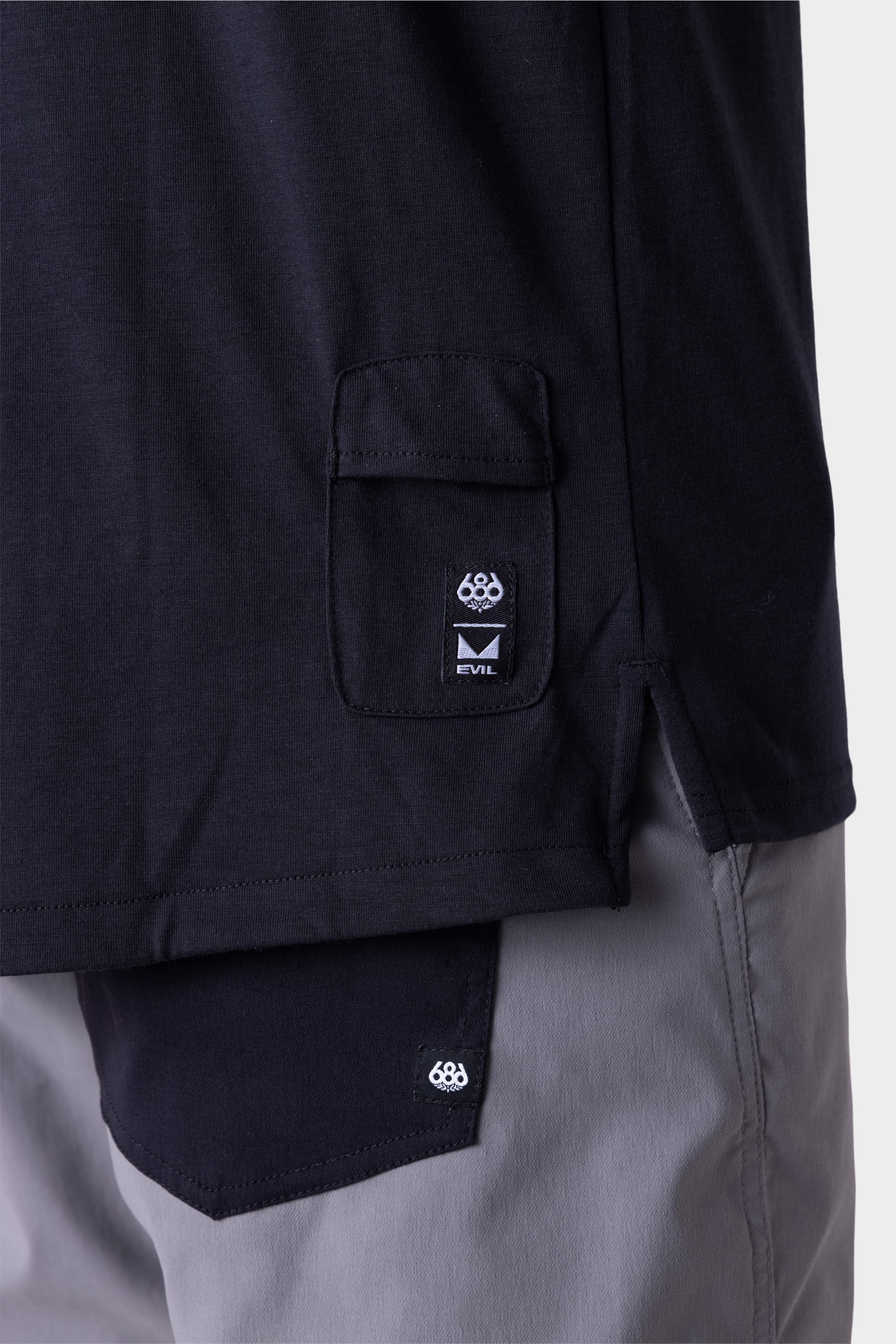 Close-up of a person wearing the 686 Mens Rival Short Sleeve Bike Jersey in black, featuring a flap pocket with the 686 logo label and EVIL. The light grey pants also display a matching 686 logo on the pocket.