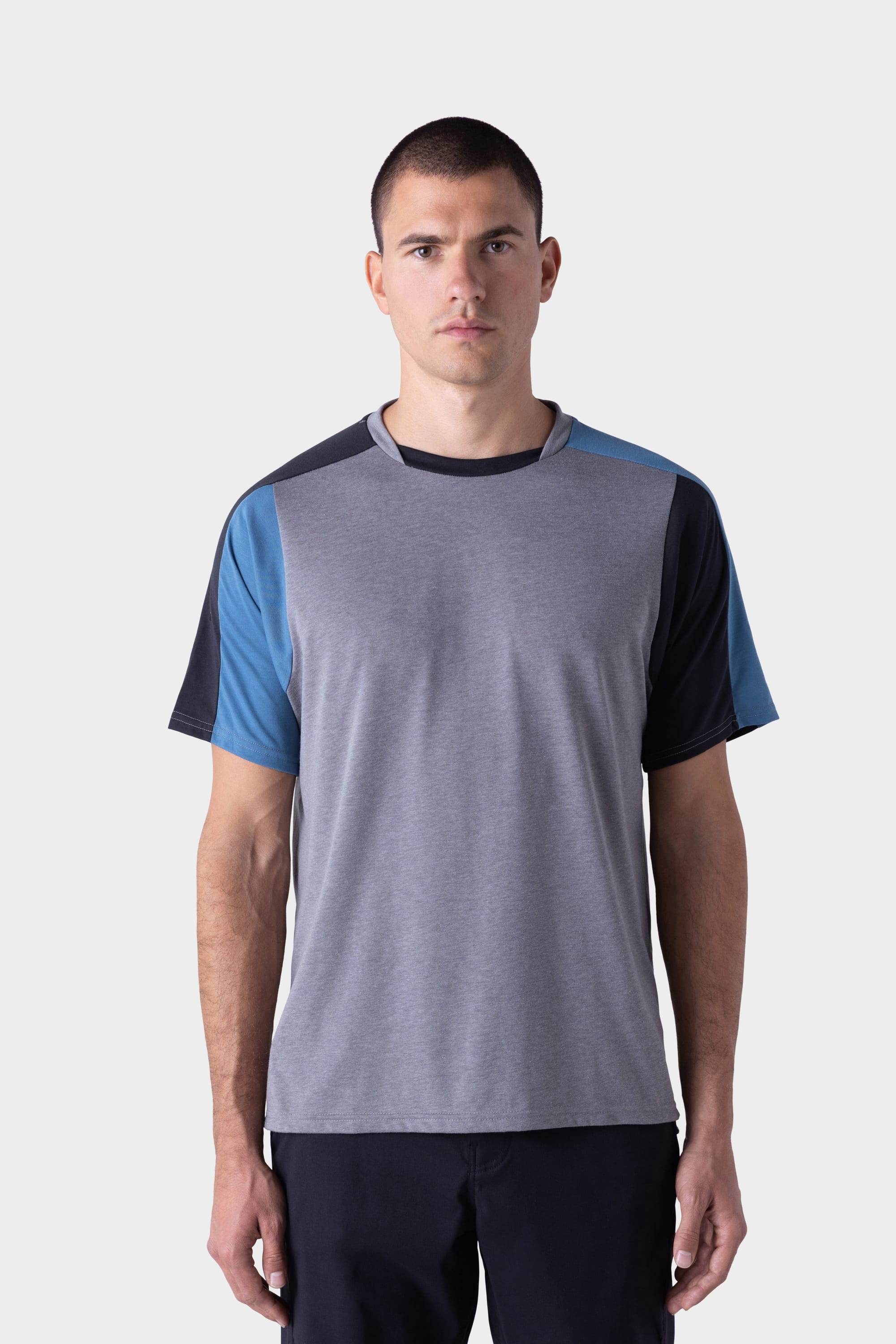 A man with short dark hair stands facing forward, wearing the 686 Mens Rival Short Sleeve Bike Jersey by 686, which features grey fabric with blue and black color-blocked sleeves, and dark pants, against a plain light background.