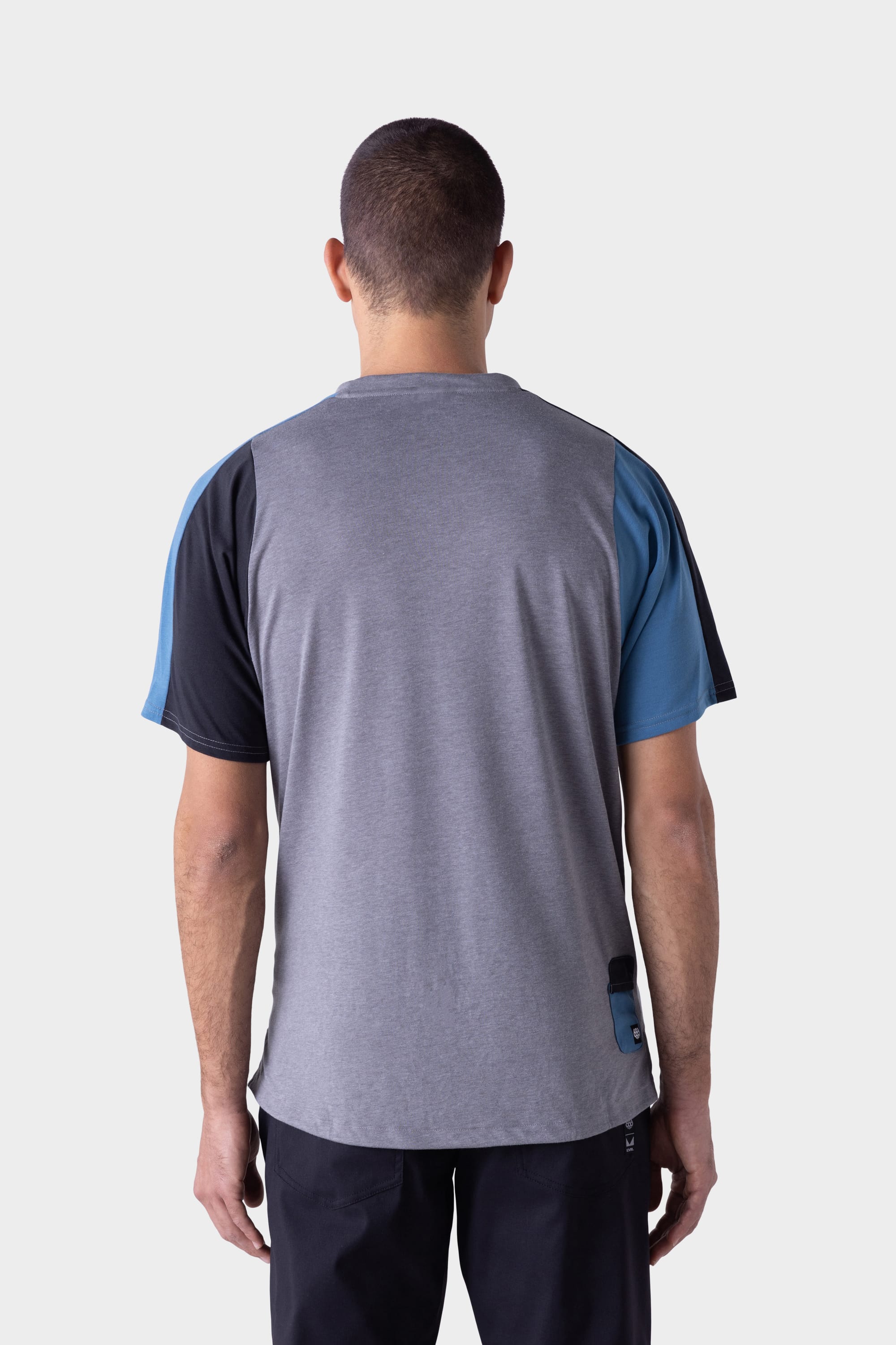 A man stands facing away from the camera, wearing the 686 Mens Rival Short Sleeve Bike Jersey with grey, black, and blue panels from 686, paired with dark pants against a plain light background.