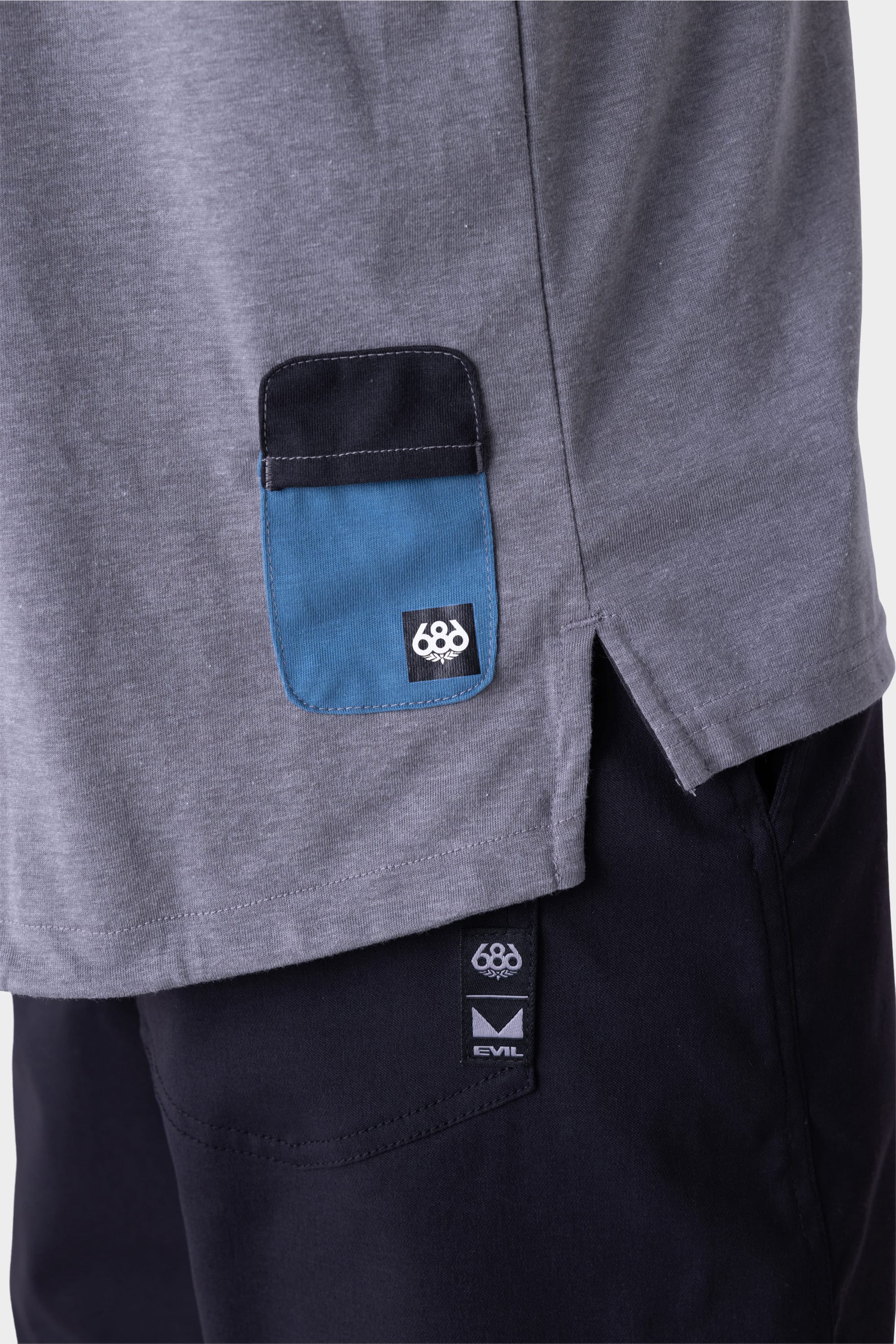 Close-up of the 686 Mens Rival Short Sleeve Bike Jersey in grey with a small blue and black pocket featuring the 686 logo, paired with black pants displaying a visible 686 EVIL tag near the waistband.