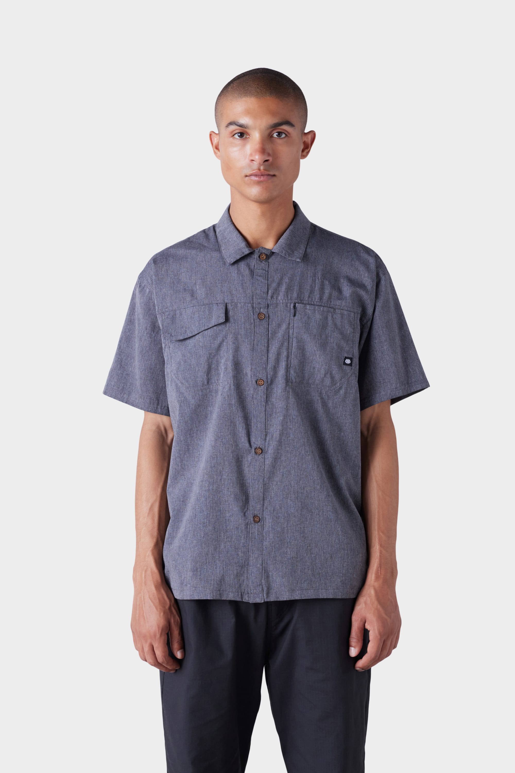 A person faces forward against a white background, wearing the 686 Mens Seeker Perforated Button Up by 686, a short-sleeve grey shirt with two chest pockets, paired with black pants.