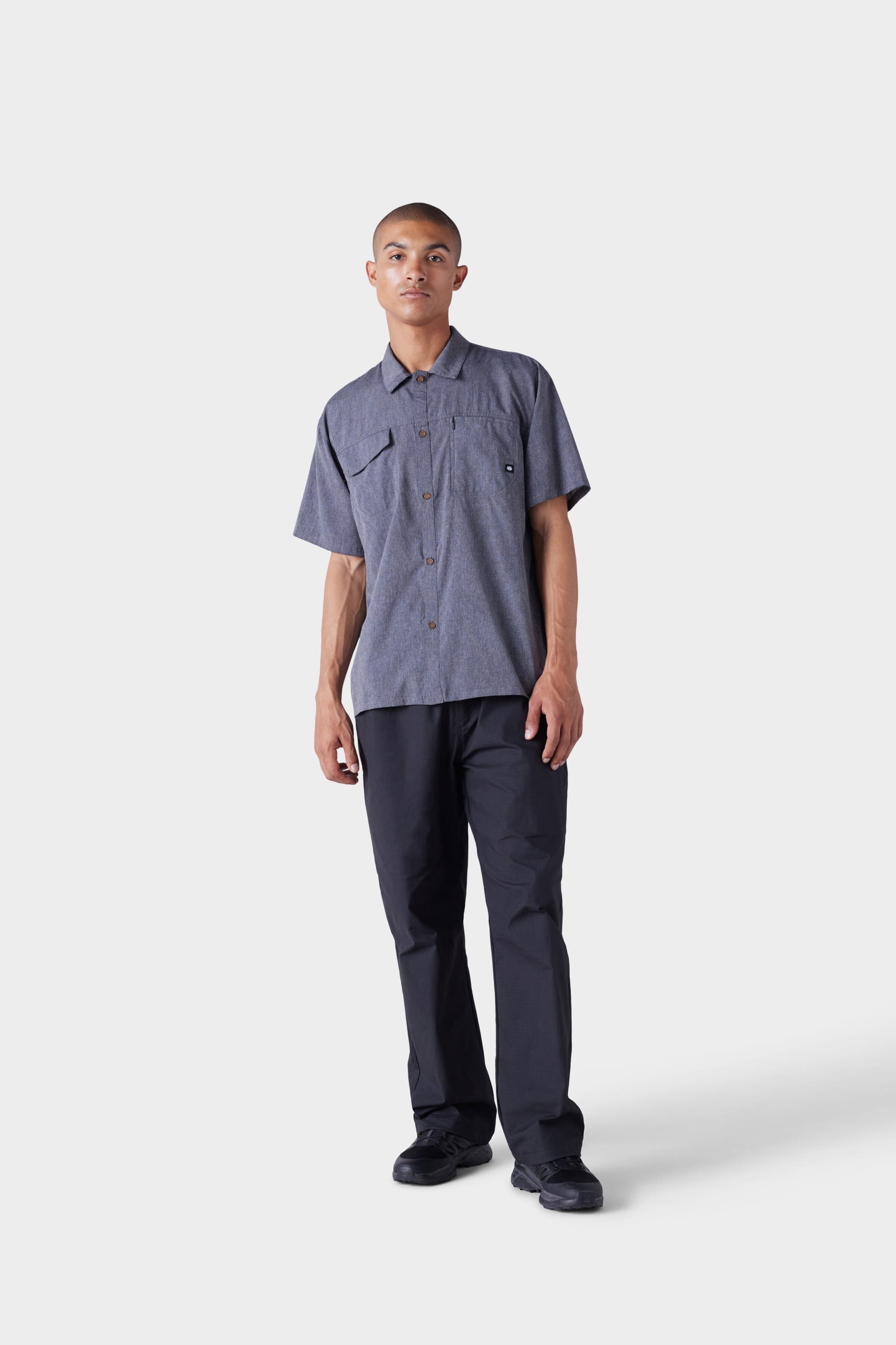 A person with short hair and a neutral expression wears the 686 Mens Seeker Perforated Button Up grey shirt by 686, featuring two chest pockets, paired with black pants and black shoes against a plain white background.