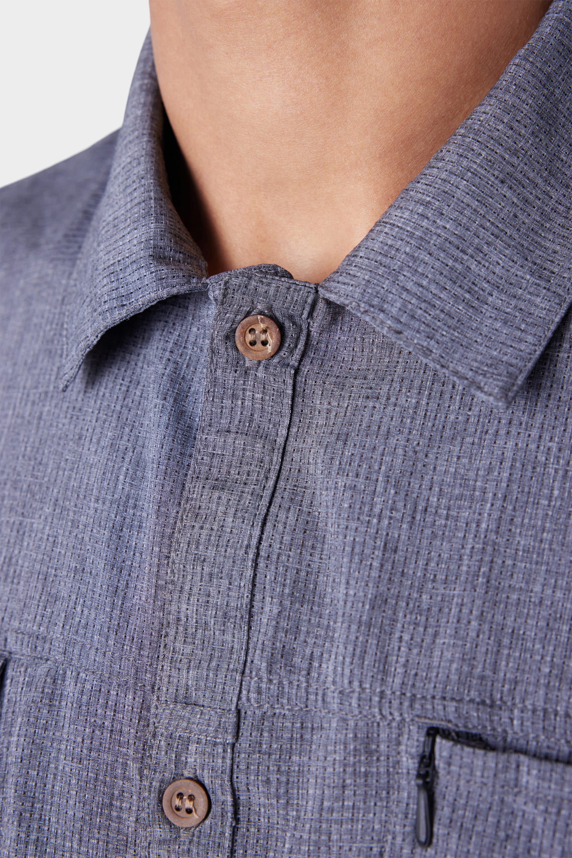 Close-up of a person wearing the 686 Mens Seeker Perforated Button Up, a textured blue-grey collared shirt by 686 with brown buttons and a visible vertical zipper pocket. The background is plain white.