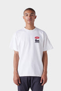 A person wearing the 686 Mens Under the Summer Sun Short Sleeve Tee, a white t-shirt by 686 with a small red burst and black graphic on the upper left chest, stands against a plain light background.
