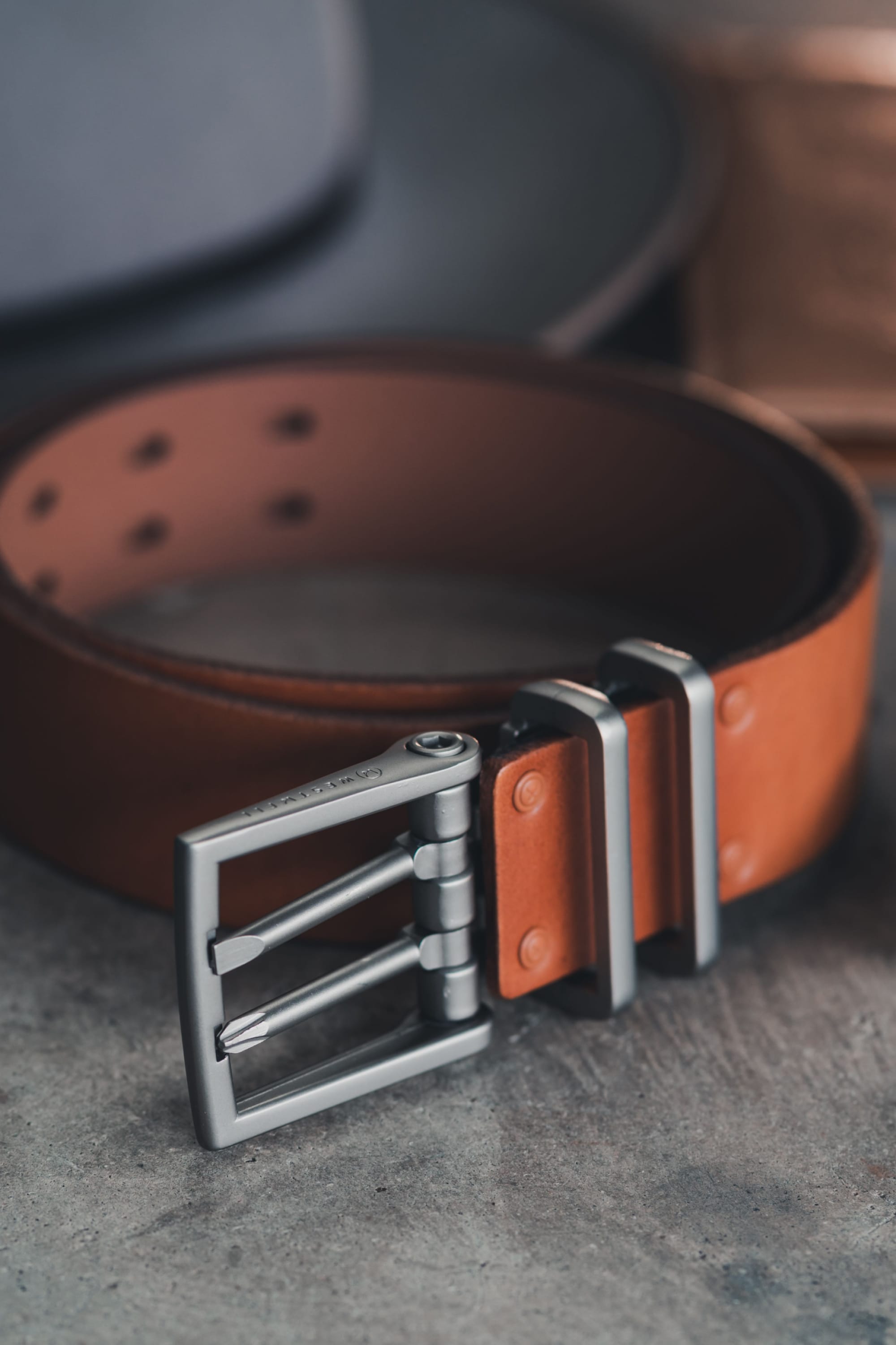 The 686 Westwell Voyager Belt by Westwell is shown up close on a gray surface, featuring brown leather, a silver metal double-prong buckle, metal loops, visible stitching, and multiple adjustment holes.
