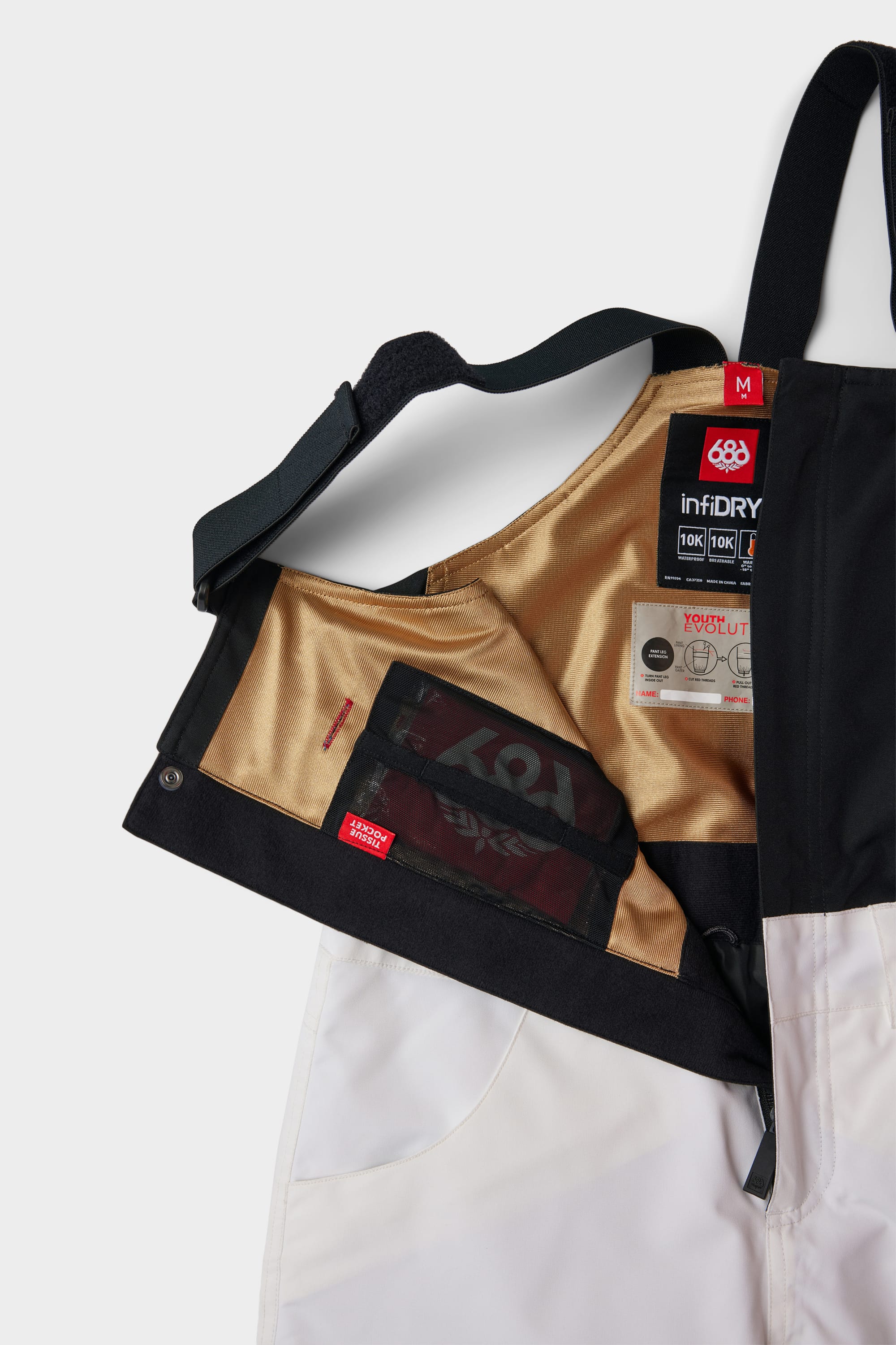 The 686 Youth Exploration Insulated Bib is shown open, revealing its gold inner lining, mesh pocket, and tags with washing instructions and 686 brand info. White snow pants feature black suspenders for a stylish contrast.