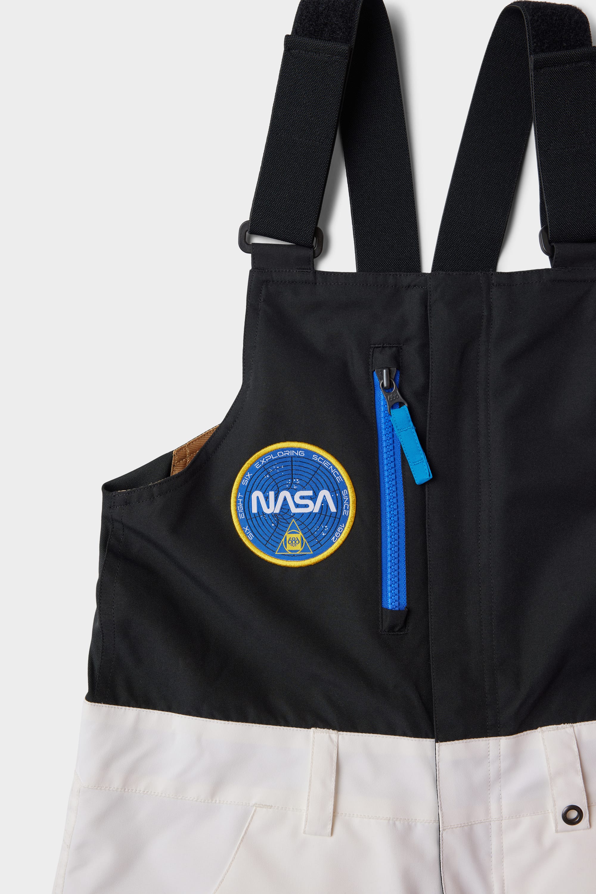 A close-up of the 686 Youth Exploration Insulated Bib by 686, featuring black adjustable straps, a front zipper pocket with a blue pull, and a round blue NASA patch on the chest area.