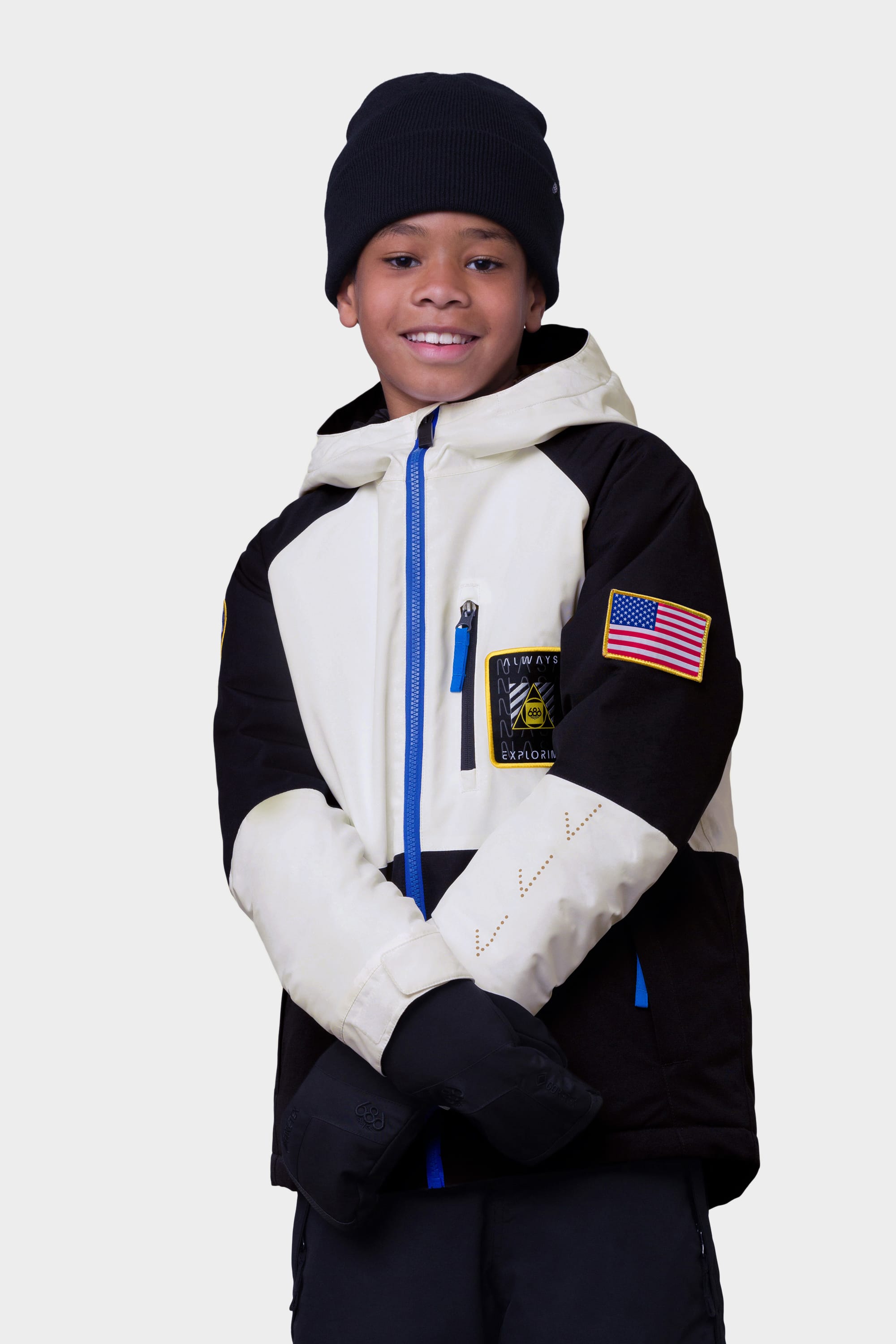 A smiling child in the 686 Youth Exploration Insulated Jacket by 686, featuring a black and white design with a U.S. flag patch, plus a black beanie and gloves, stands against a plain light background.