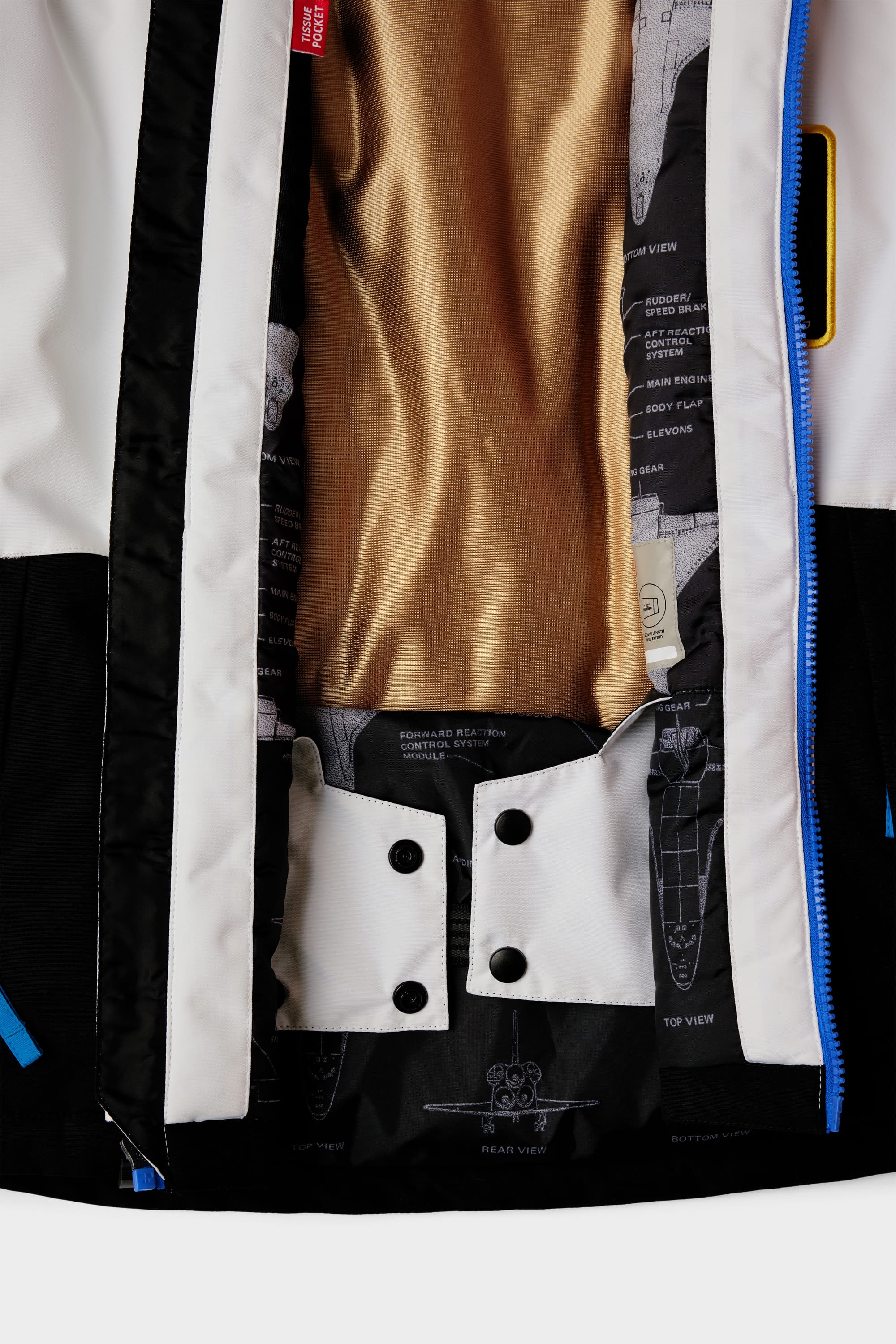 Close-up of the 686 Youth Exploration Insulated Jacket by 686, featuring a gold inner lining, black and white panels, blue zipper, and an inner label with robot graphics. Two black buttons are fastened on a white flap.