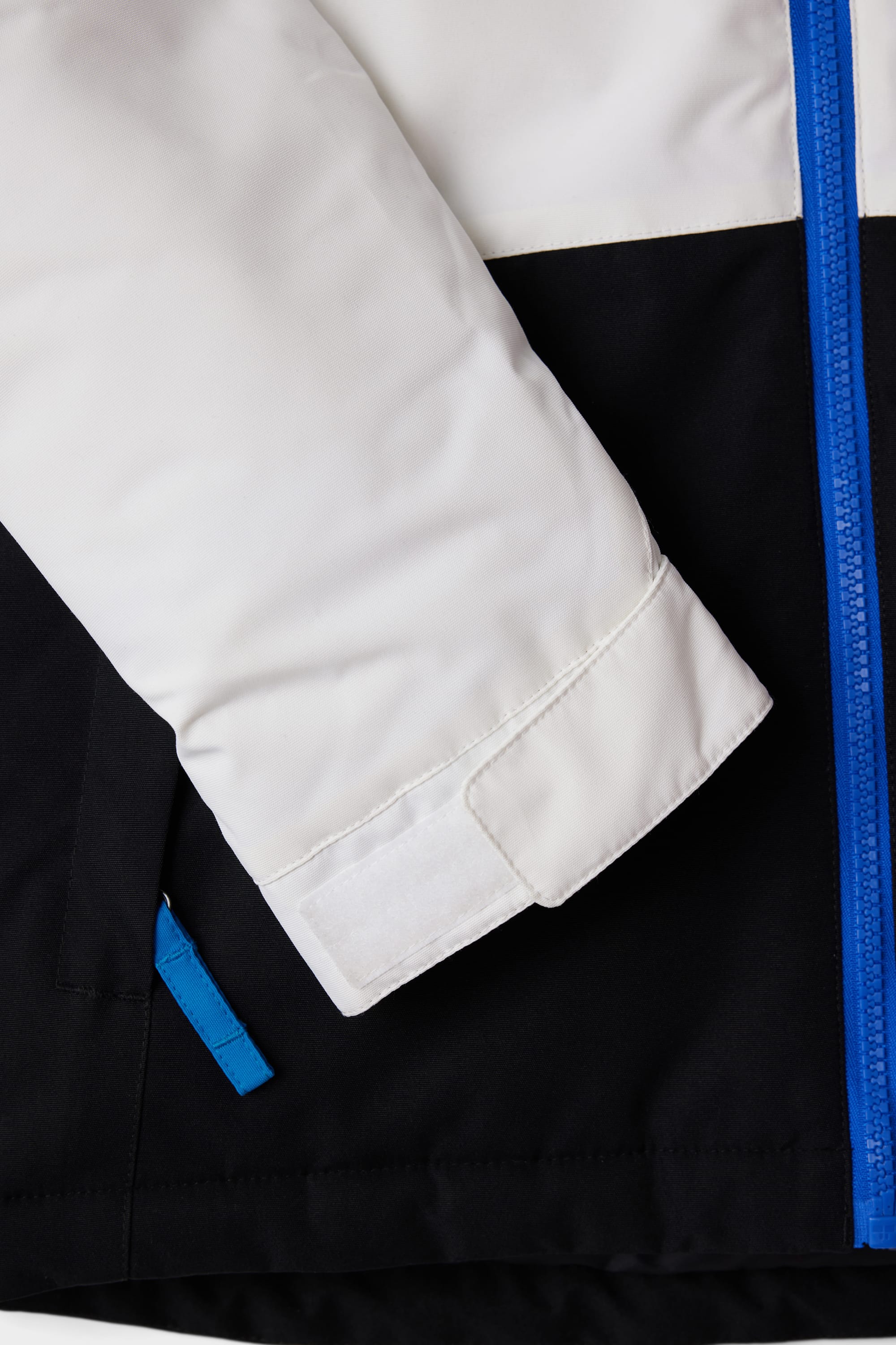 Close-up of the 686 Youth Exploration Insulated Jacket sleeve shows a white upper with Velcro strap, blue zipper, and black lower section featuring a pocket with blue pull tab.