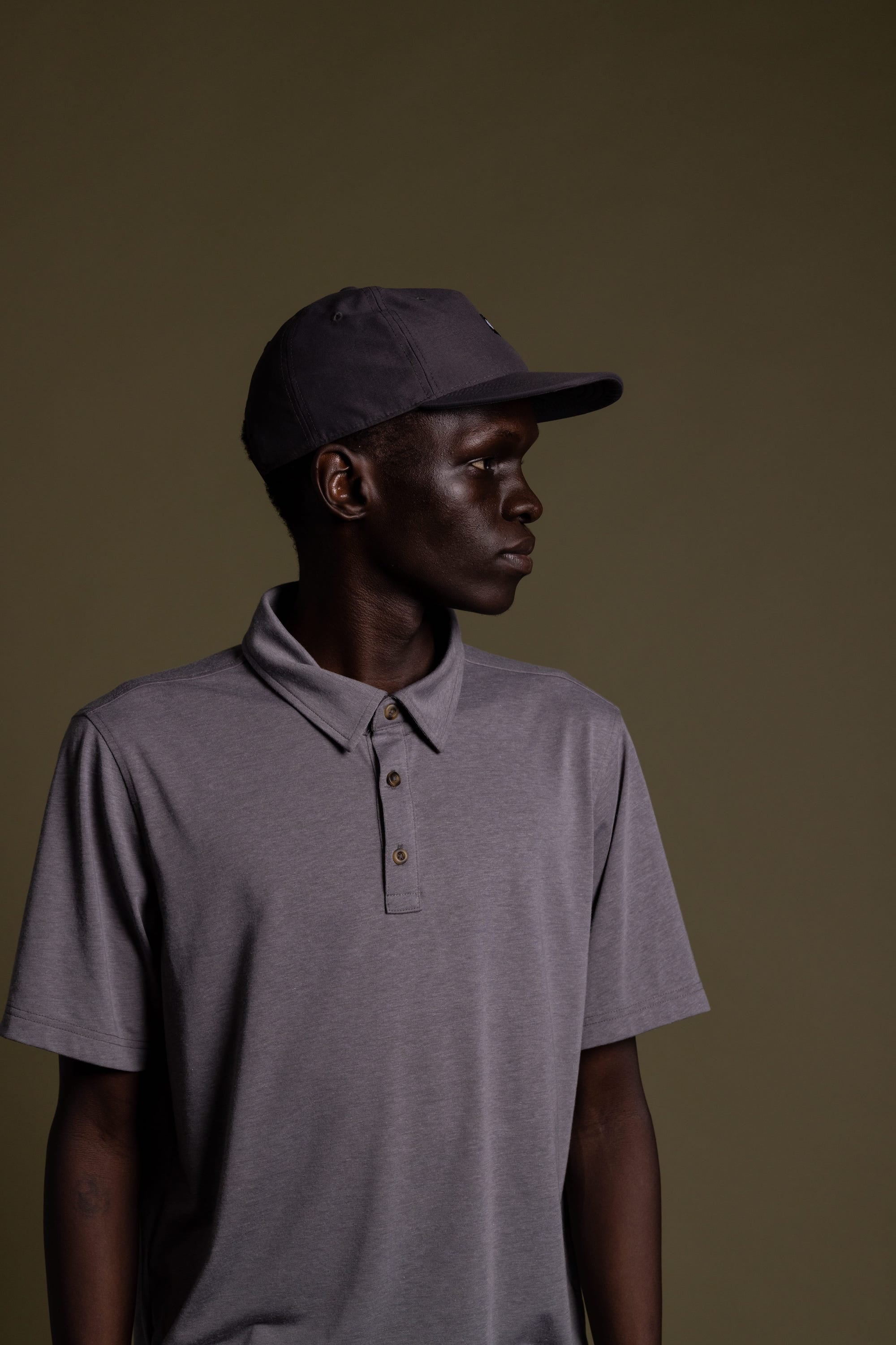 A person wearing a gray 686 Packable Everywhere Hat by 686 stands against a plain olive-green background, looking to the left with a neutral expression.