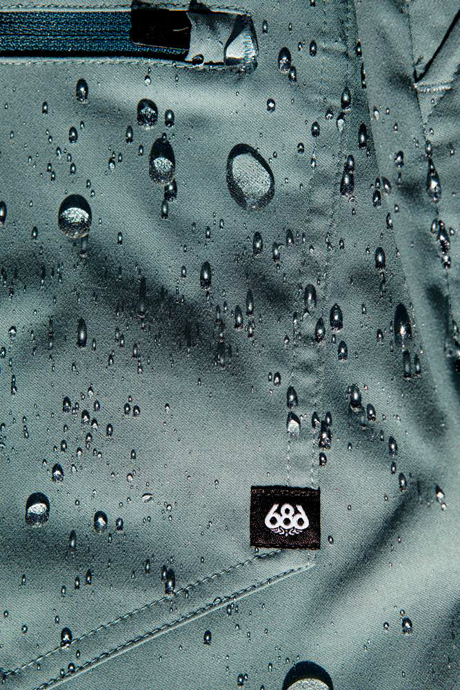 Close-up of water droplets on teal, stain-resistant, breathable fabric with a zipper and black 686 tag, showcasing the durability of the 686 Mens Everywhere Hybrid Short.