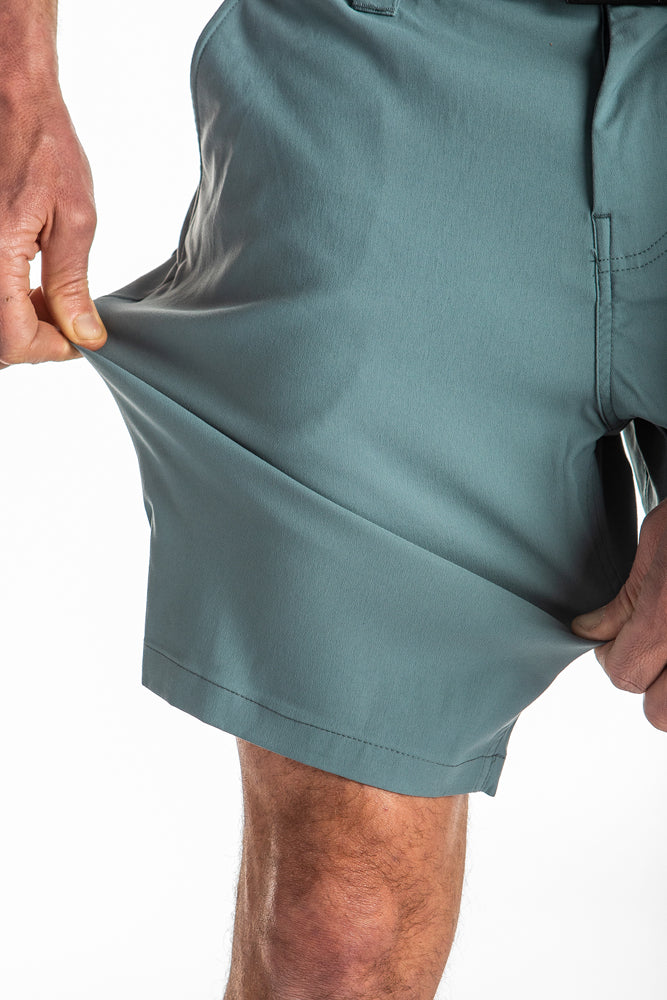 A person stretches the 686 Mens Everywhere Hybrid Shorts light blue-green fabric with one hand to show its flexibility. The image is cropped to display only the shorts and the persons hands.
