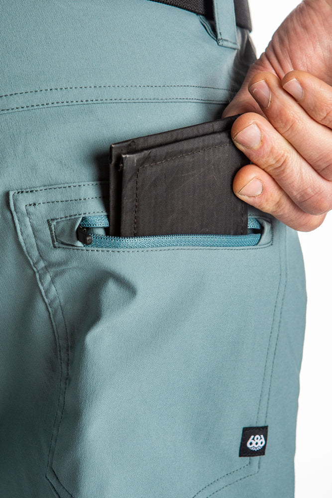 A hand places a black wallet into the zippered back pocket of light blue, breathable 686 Mens Everywhere Hybrid Shorts.