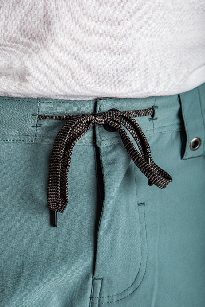 Close-up of a person wearing 686 Mens Everywhere Hybrid Short in breathable teal, featuring a black and white drawstring bow at the waist, paired with a white shirt partially tucked in.
