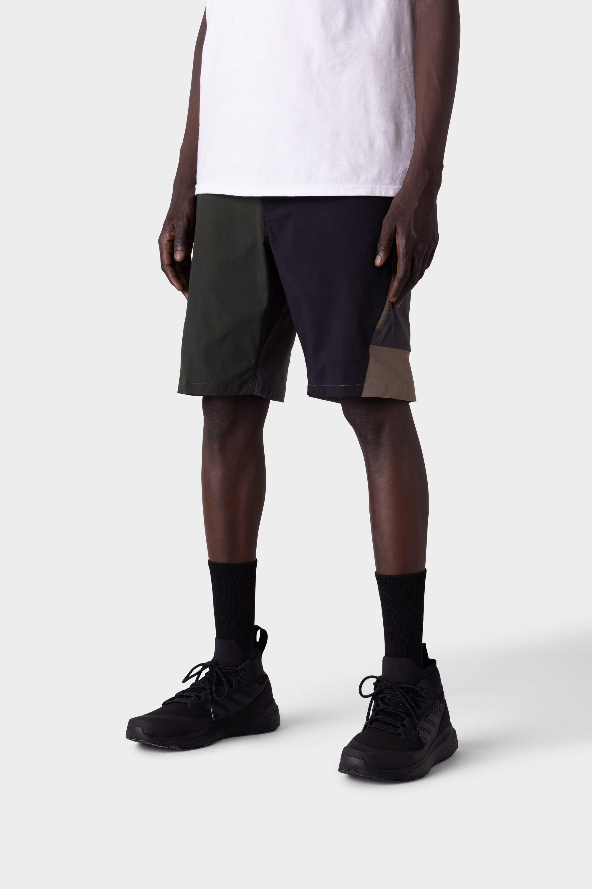 Someone stands against a plain light gray background wearing a white t-shirt, 686 Mens Anything Hybrid Cargo Short by 686 in color-blocked black, olive, and brown, paired with black crew socks and black sneakers.
