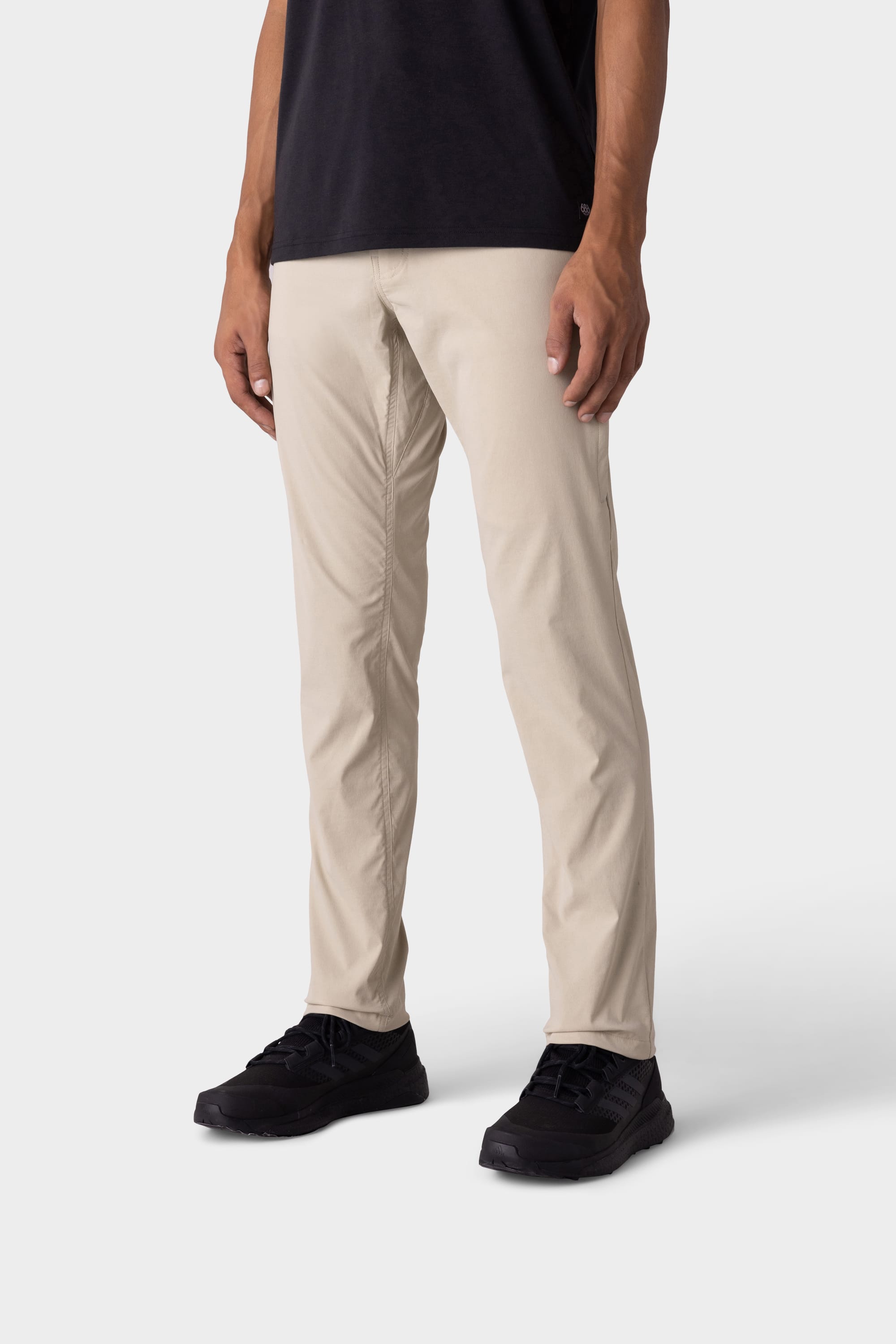 A person wears 686 Mens Everywhere Featherlight Chino Pant - Slim Fit in beige, paired with a black t-shirt and black sneakers, standing against a light grey background—a perfect choice from 686 for your summer wardrobe and warm weather outings.