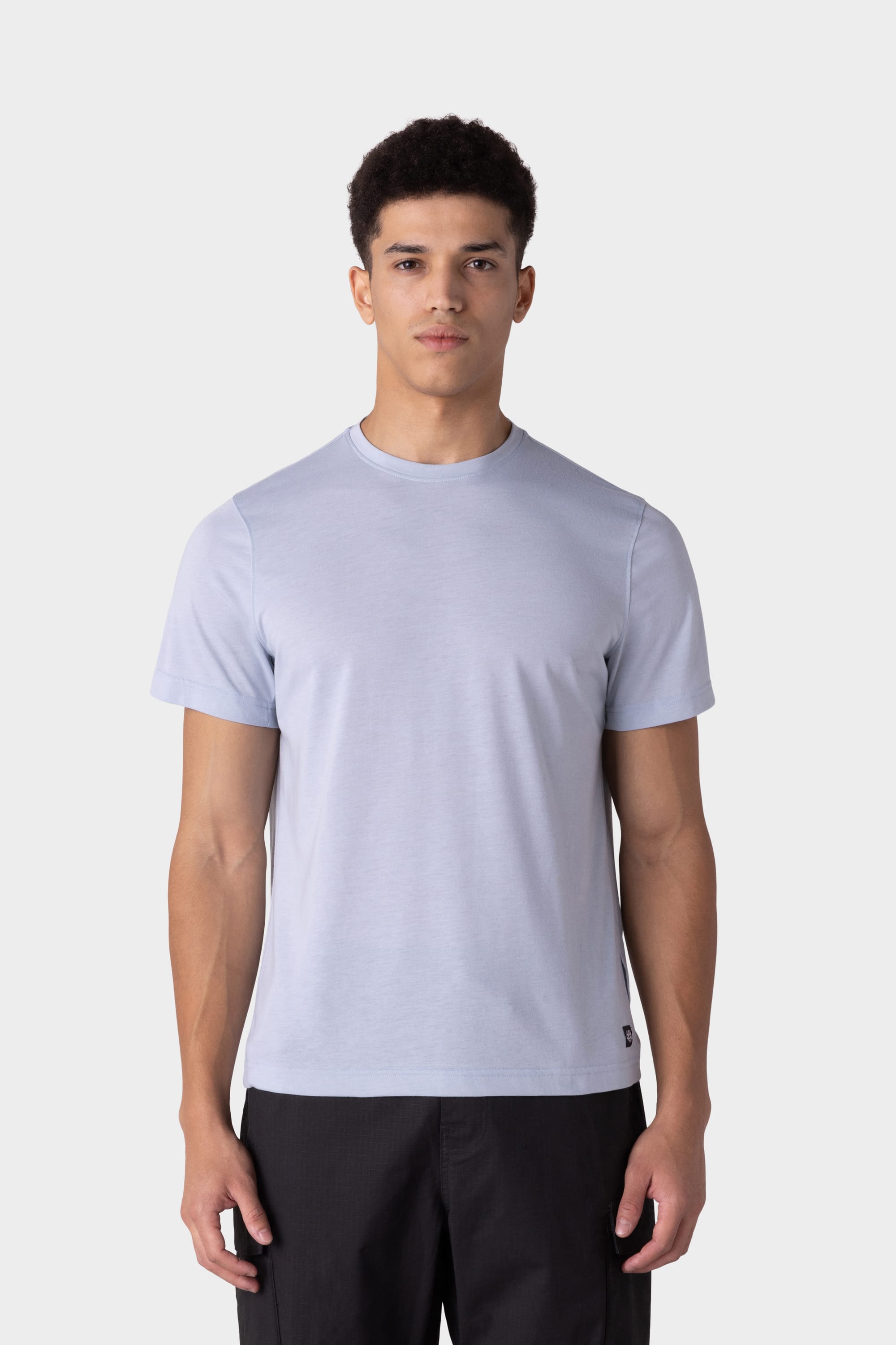 A young man with short curly hair models the 686 Mens Everywhere drirelease® Tech Tee in light gray with black pants, standing against a plain white background and facing forward with a neutral expression.