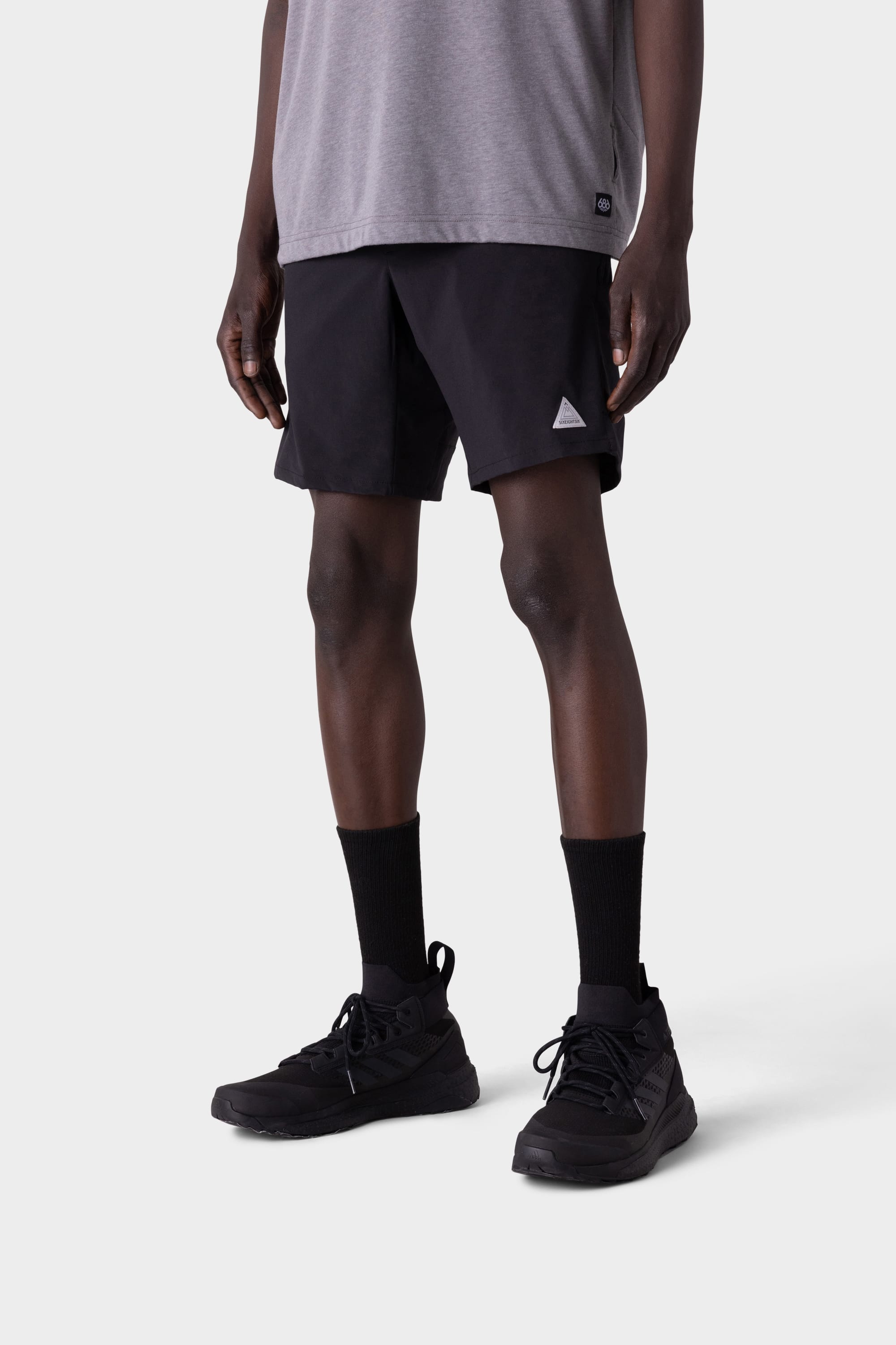 A person wearing 686 Mens ATP Stretch Performance Short by 686, black crew socks, and black sneakers is standing against a light grey background. Only the lower body is visible.