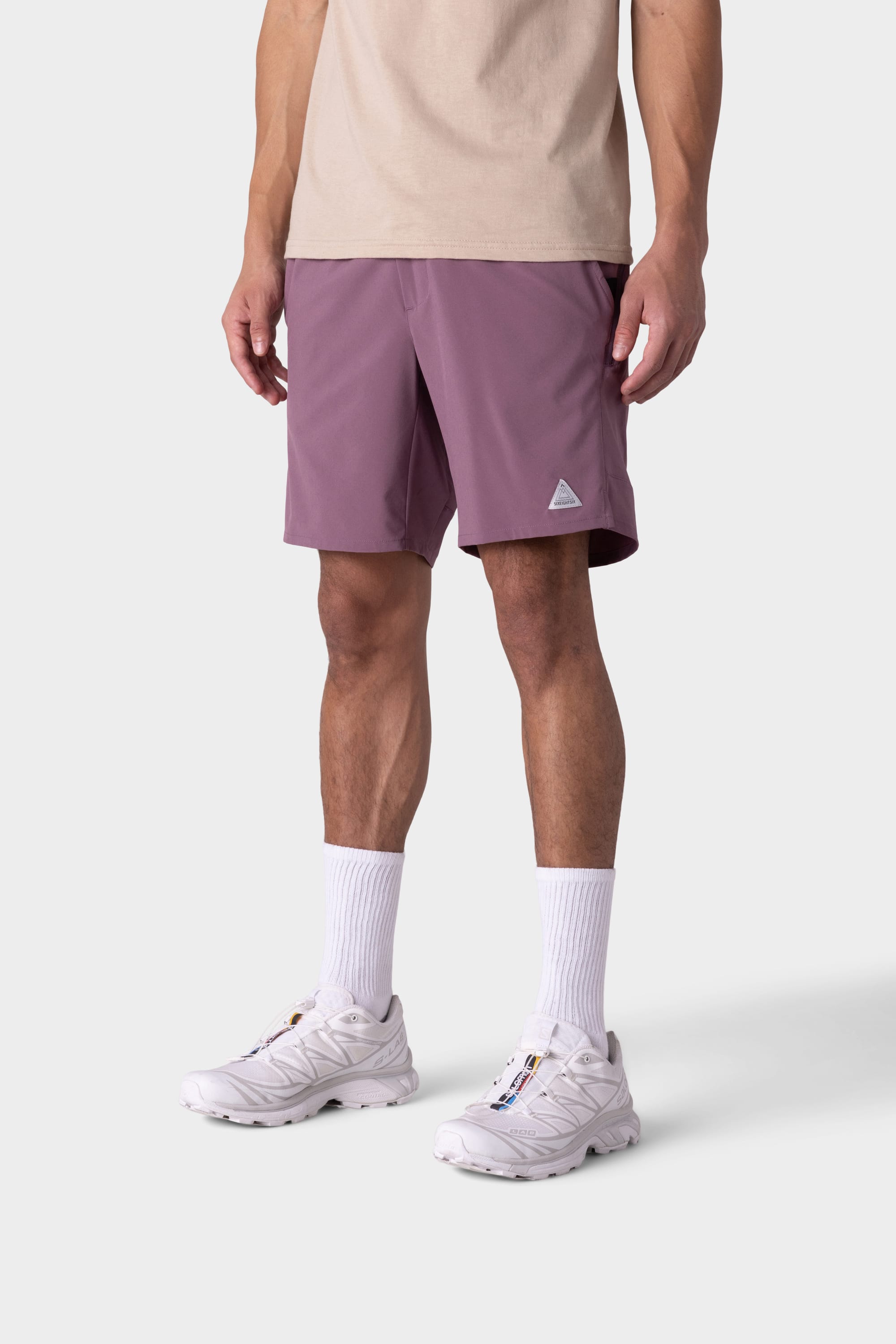 A person wearing 686 Mens ATP Stretch Performance Short in mauve, a beige shirt, white crew socks, and white sneakers stands against a plain light gray background; the face is not visible.