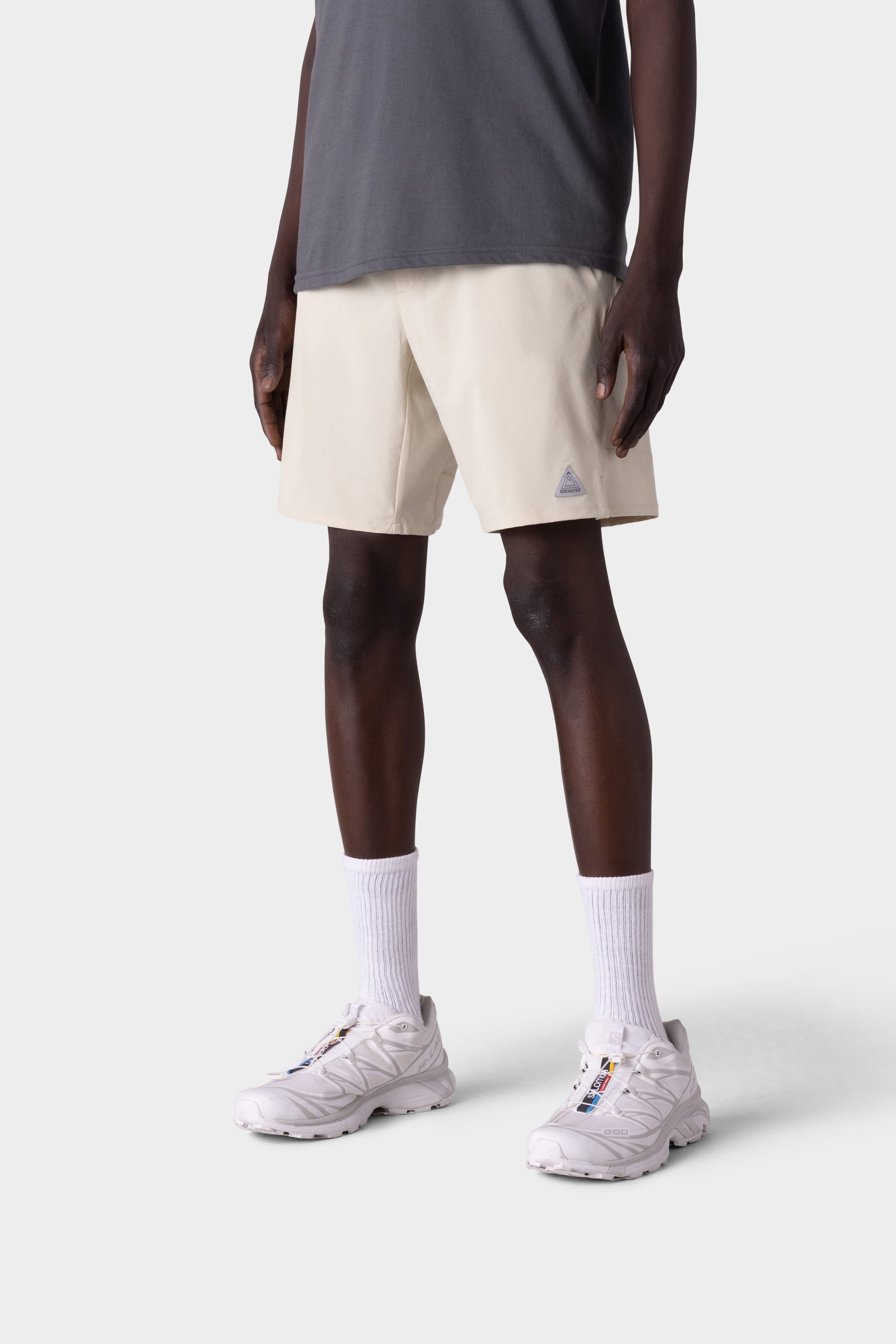 A person wearing a dark gray t-shirt, 686 Mens ATP Stretch Performance Shorts in light beige, white crew socks, and white running shoes stands against a plain white background. The image is shown from the shoulders down.